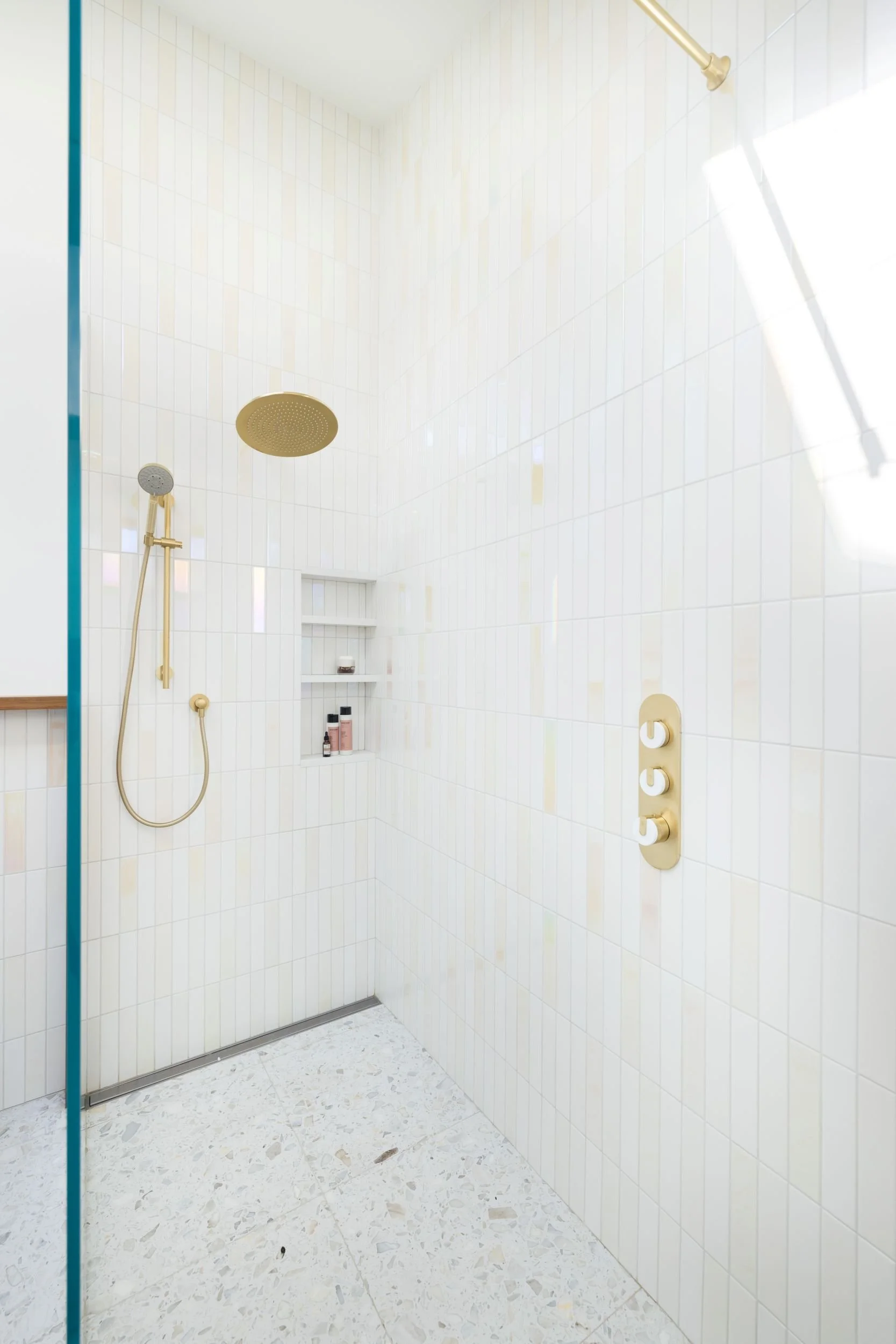 Empty shower with gold fixtures, a large overhead shower head, a handheld shower head, and a niche with toiletries on the tiled wall.