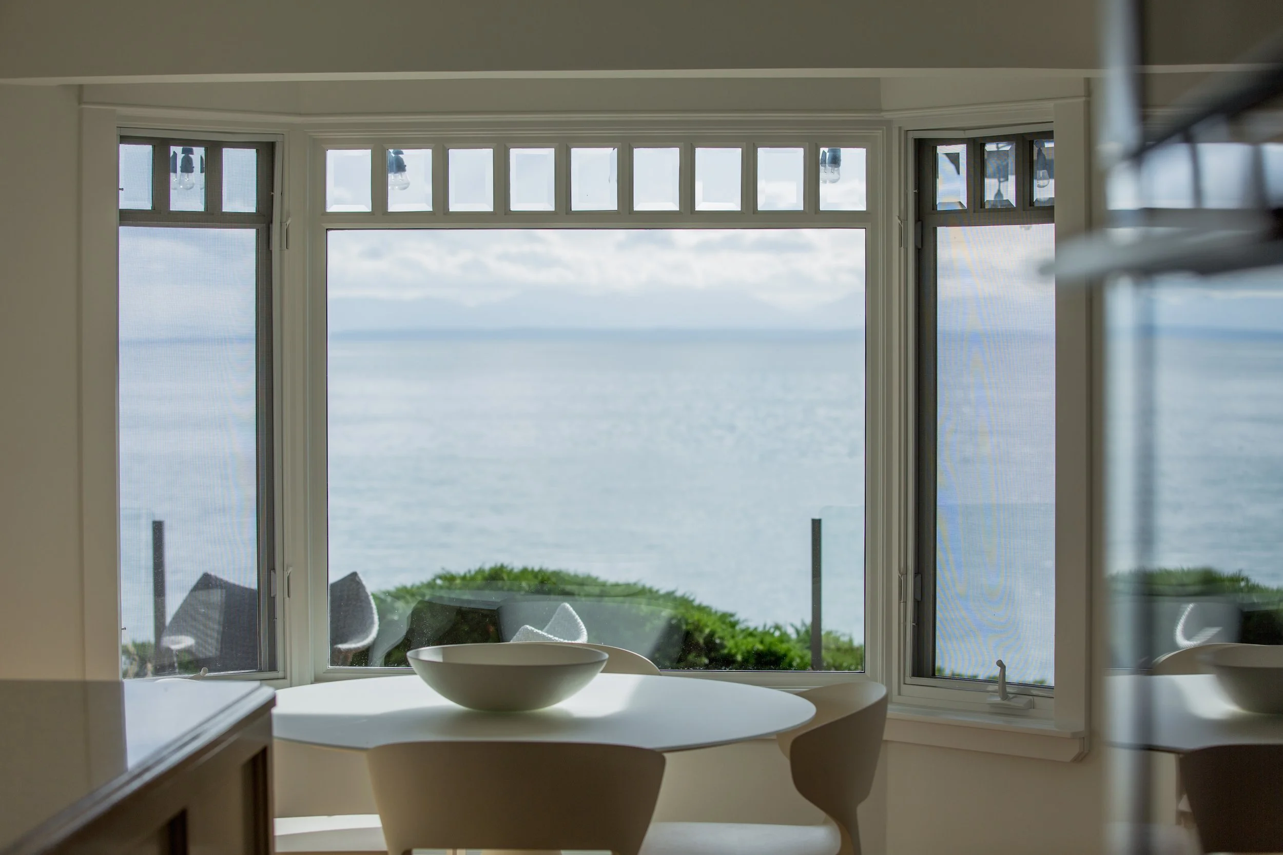 Interior view of a room with a circular white table and a bowl on top, overlooking an ocean through large bay windows with a view of the water, cloudy sky, and nearby greenery outside.