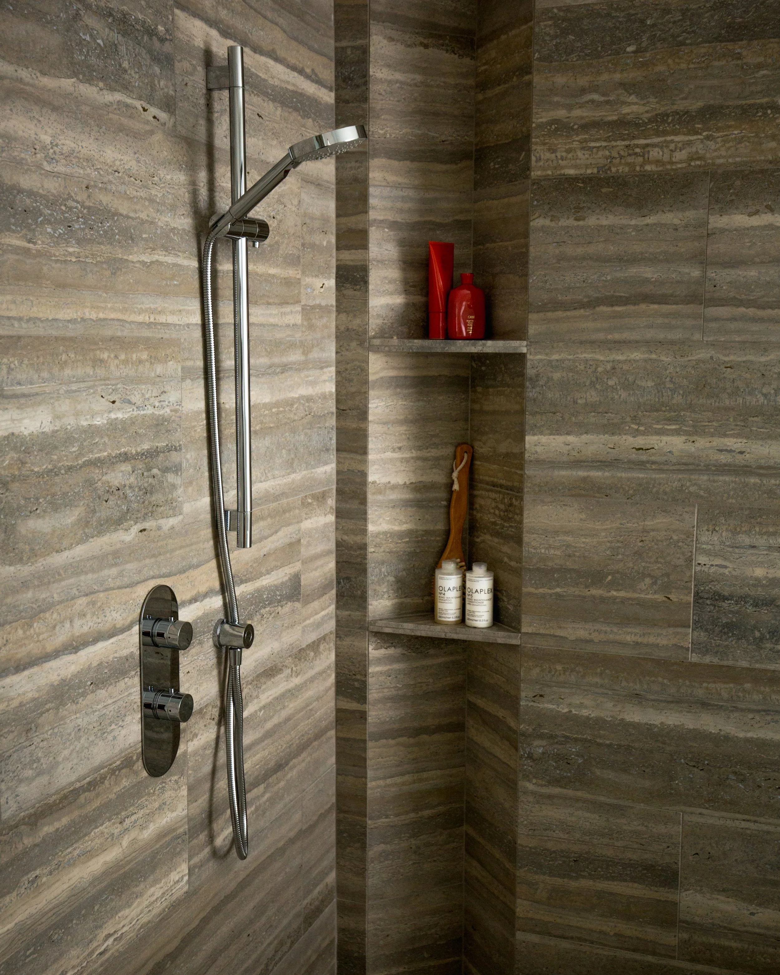 A modern shower area with gray stone tiles, a chrome shower head with a flexible hose, and a built-in shelf with red toiletries and white bottles.