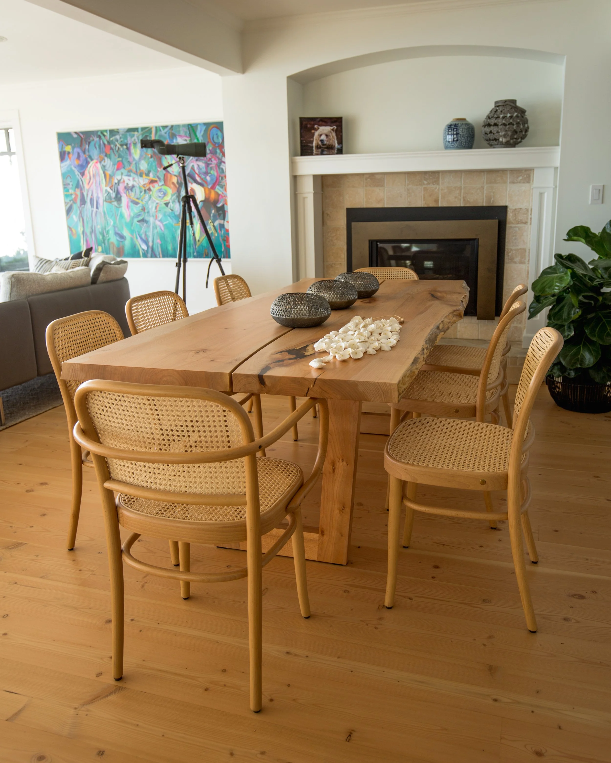Wood dining table with woven rattan chairs, decorative vases, and shell centerpiece in a living room with a fireplace, artwork, and telescope.