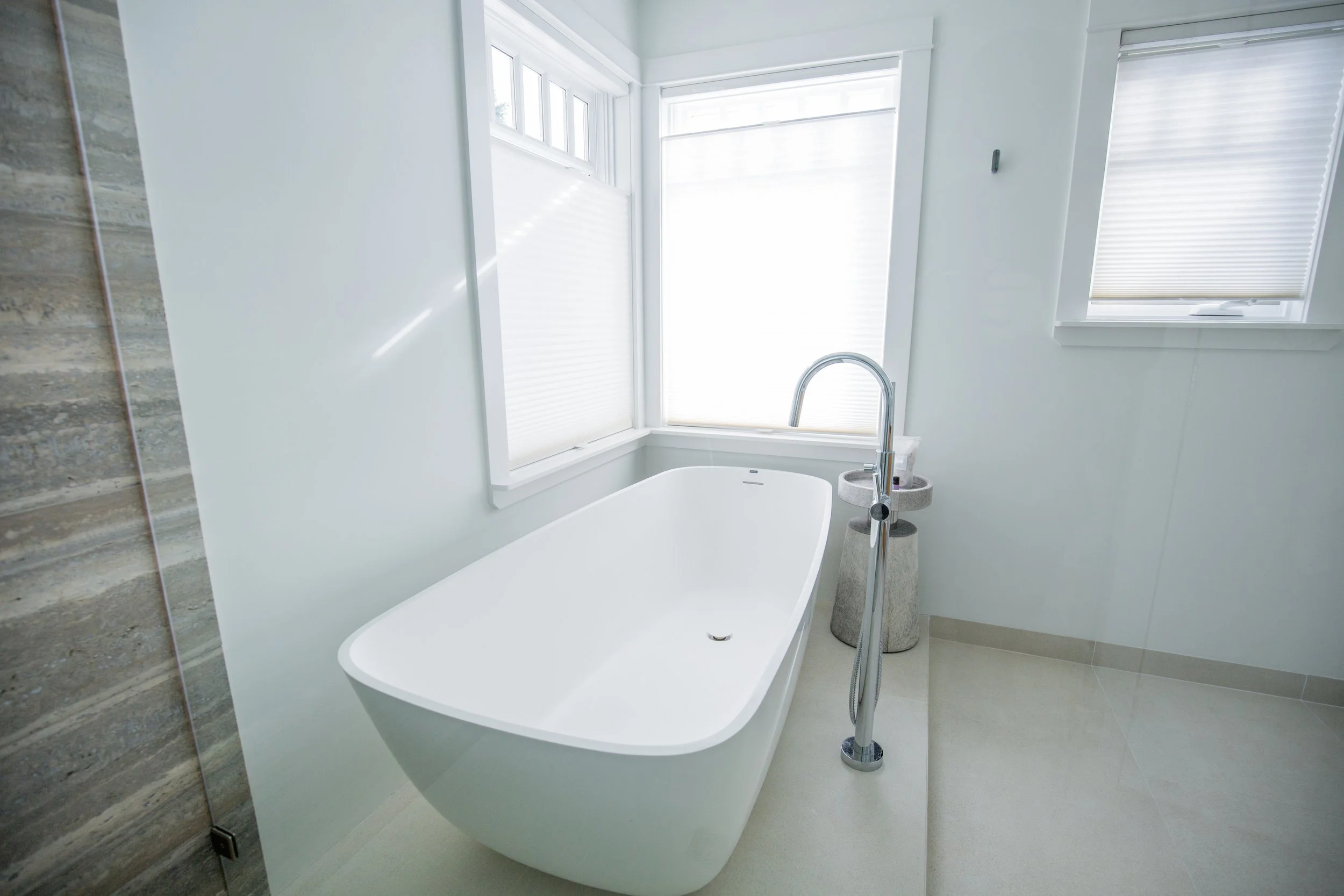 Modern bathroom with a white freestanding bathtub next to large windows with blinds, and a small table with toiletries.