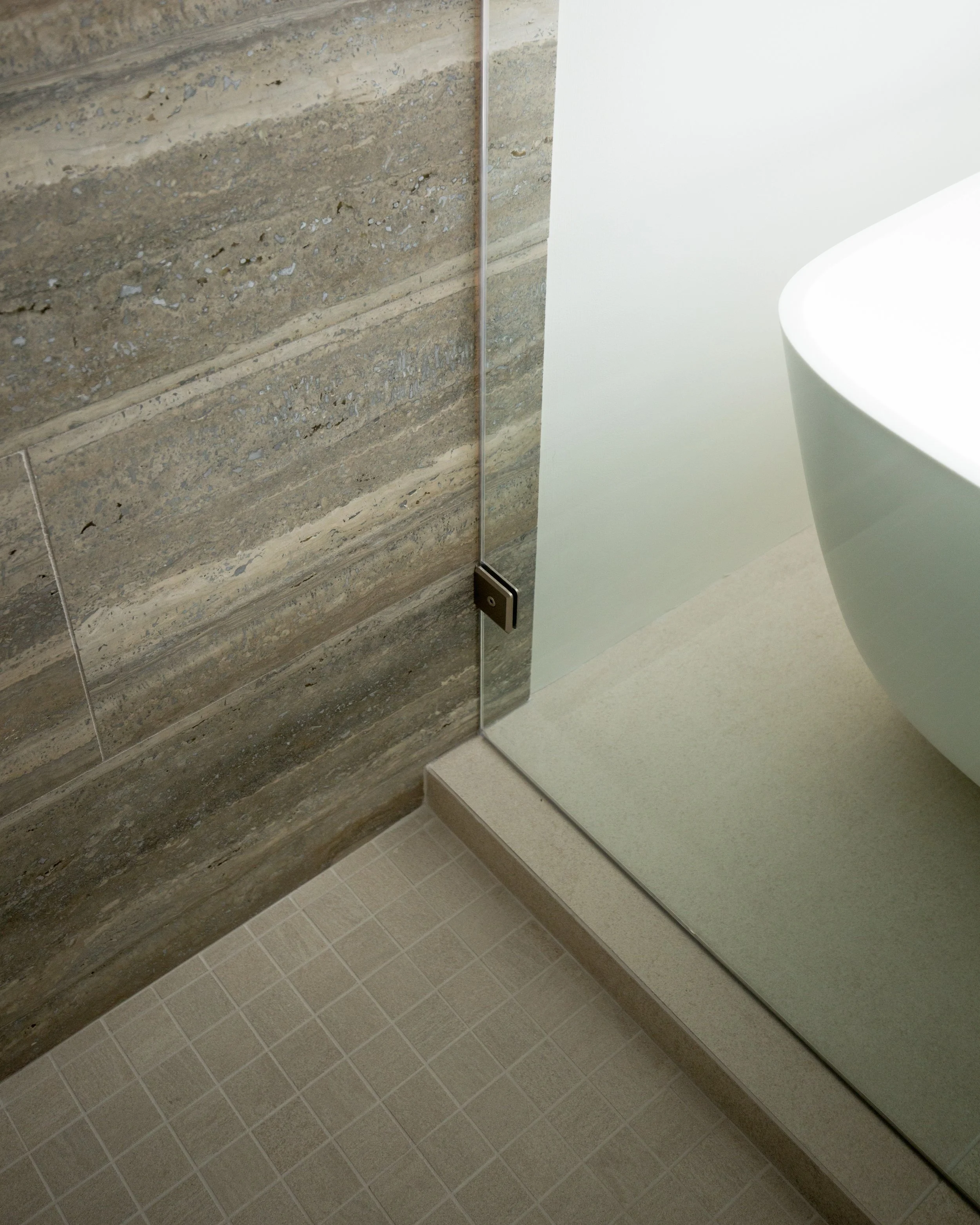 Close-up view of a bathroom corner with a textured stone wall, a glass shower enclosure with a black hinge, beige tiled floor, and a white bathtub.