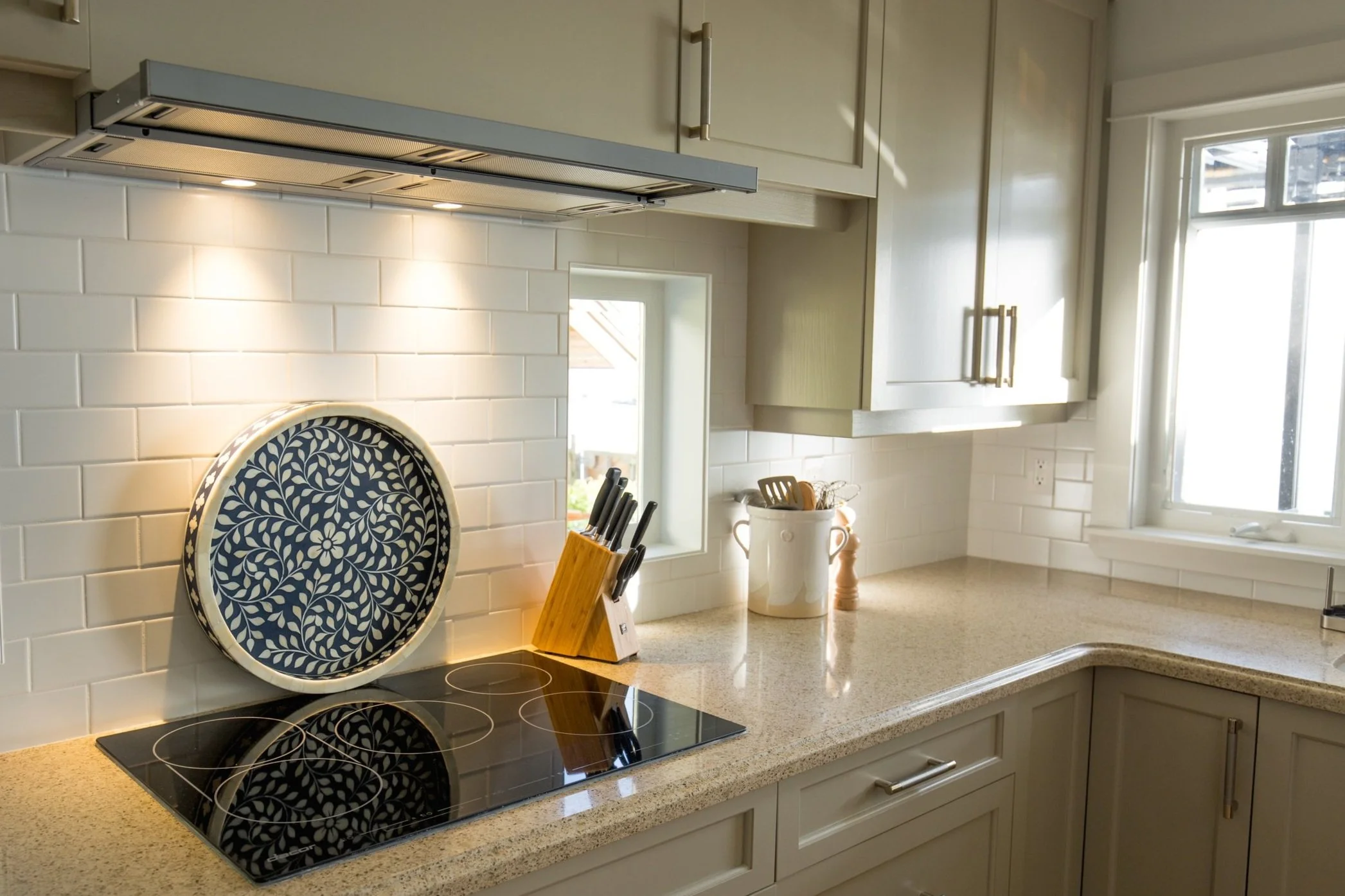 Kitchen with white cabinets, beige countertops, white subway tile backsplash, black electric cooktop, knife block, white utensil holder, and window with natural light.