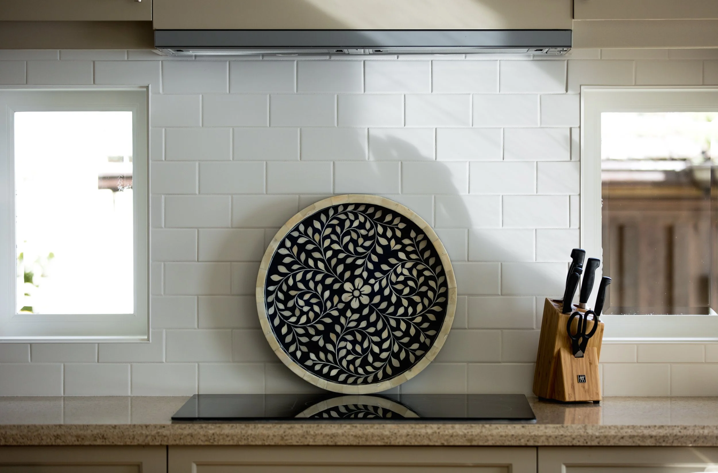 Kitchen with white tiled backsplash, black and beige decorative plate, knife set in a wooden block, and two windows with views of outdoor greenery.