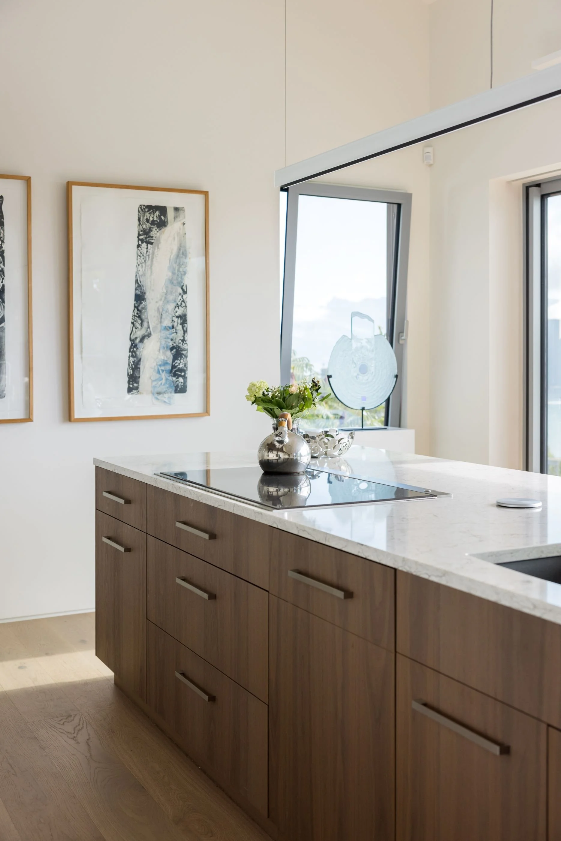 Modern kitchen with wooden cabinets, marble countertop, houseplant, and abstract wall art, with windows.
