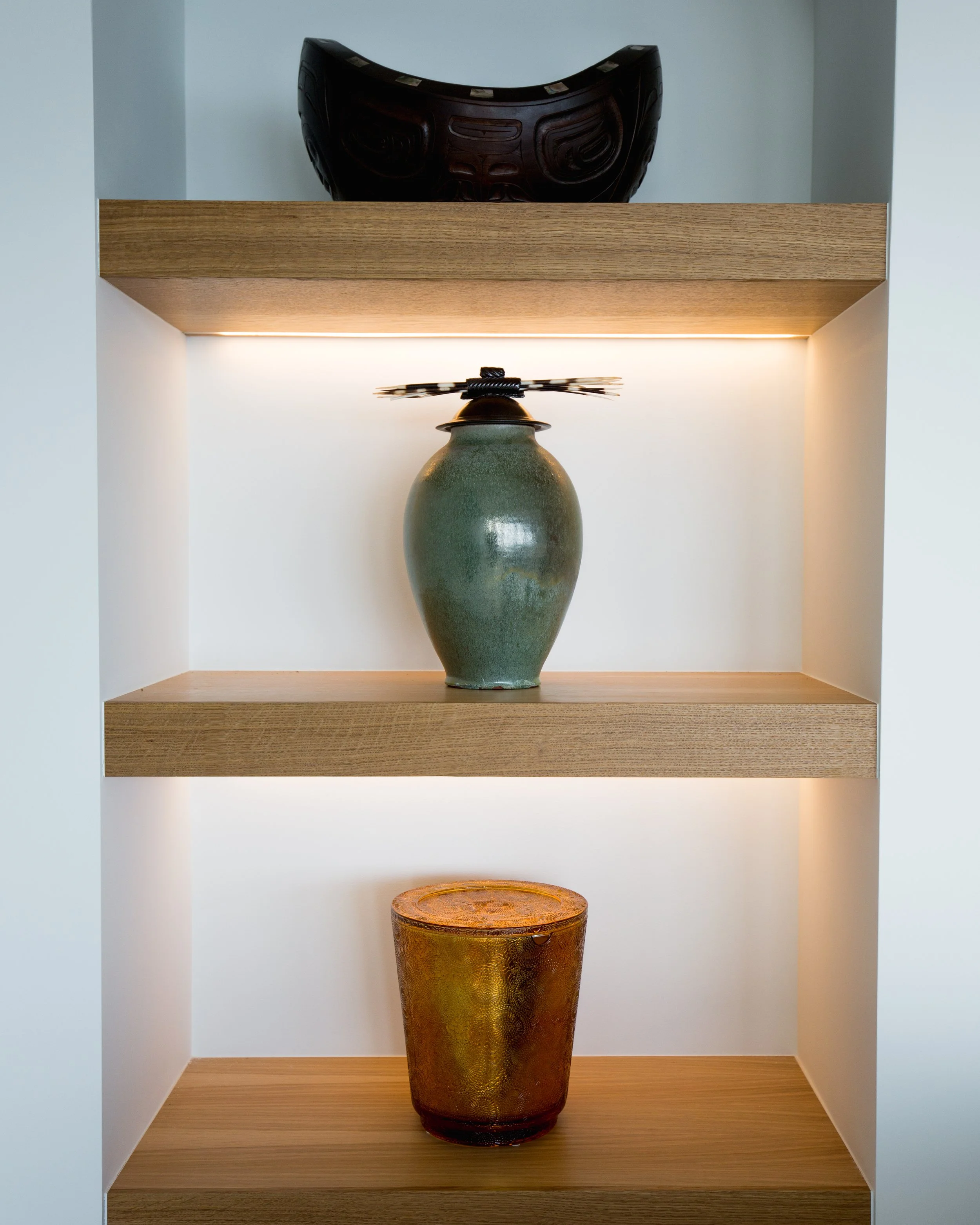 Decorative display shelf with three items: a black carved mask on top, a green ceramic vase in the middle, and a textured amber-colored glass container at the bottom.