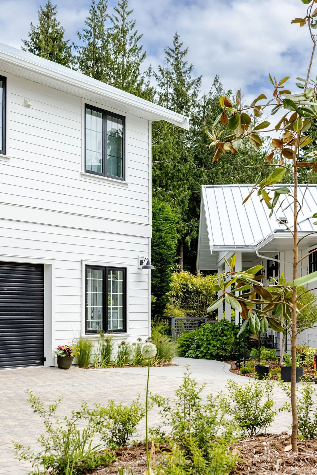 A modern white house with black window frames and a black garage door, surrounded by lush greenery and a landscaped garden with potted plants and trees.