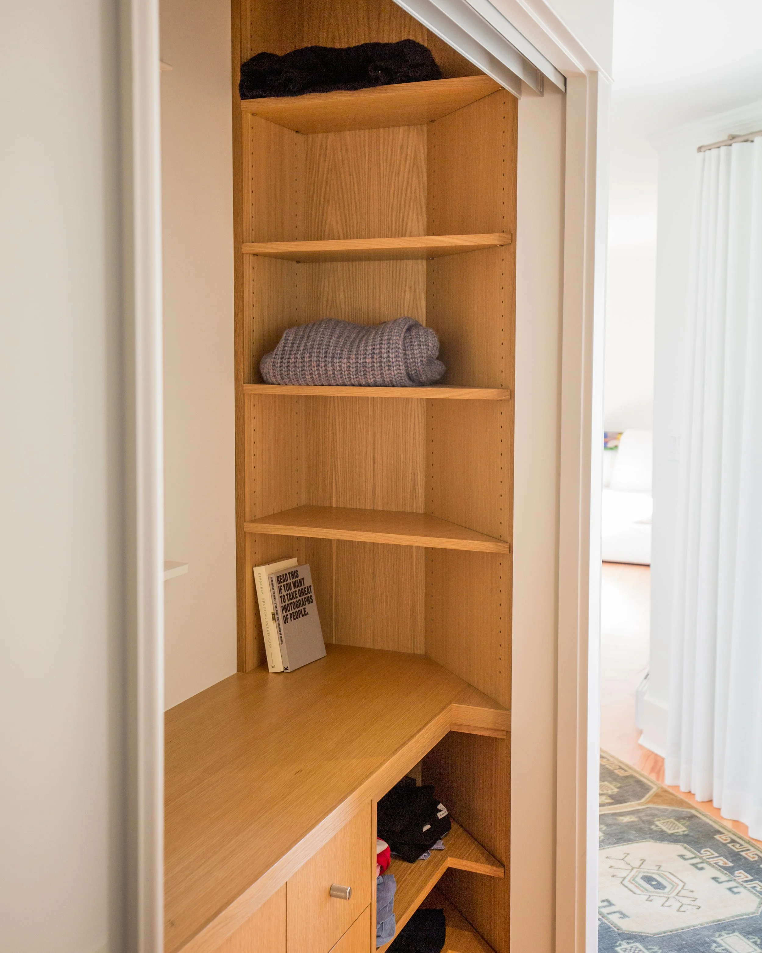 Wooden corner bookshelf with a few folded blankets, books, and miscellaneous items in a room with white walls and a rug.