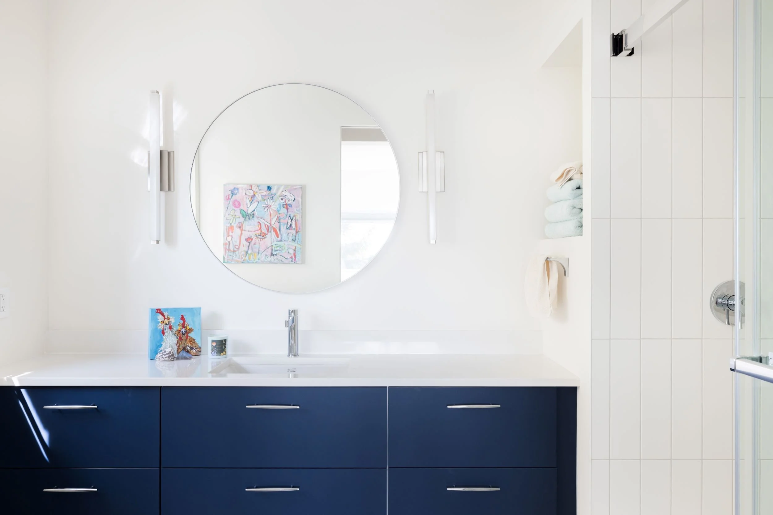 Modern bathroom with navy vanity, white countertop, and round mirror. Art on countertop and white shower with folded towels on wall.