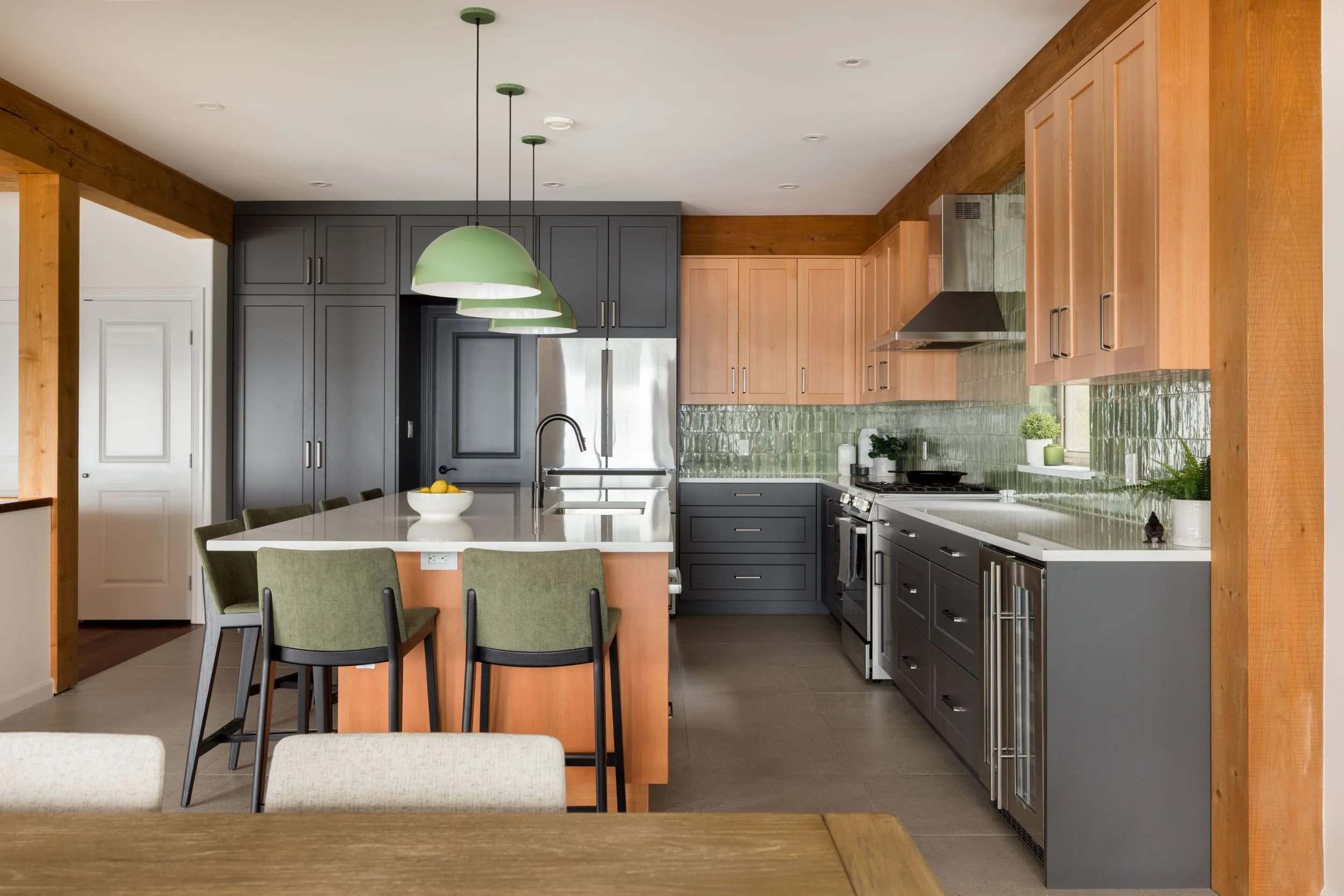 Modern kitchen with green and brown cabinets, a white island with green chairs, green pendant lights, stainless steel appliances, green tile backsplash, and wooden accents.