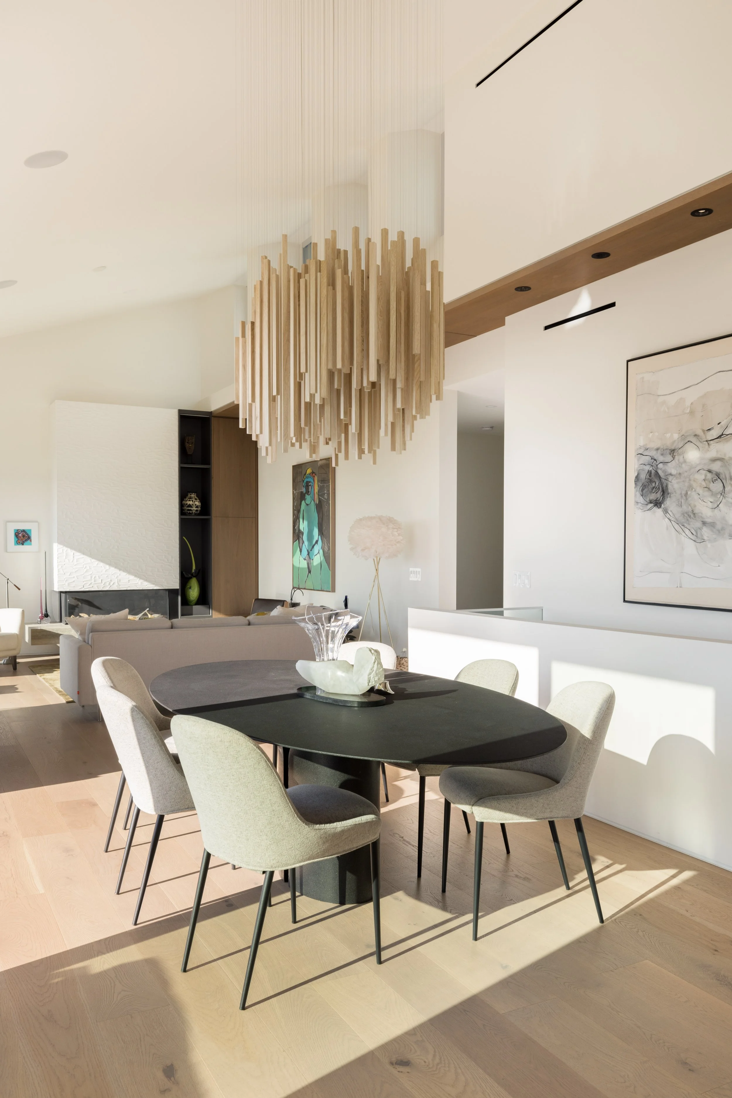 Modern dining room with black oval table, six light beige chairs, wooden chandelier, and artwork on white walls.