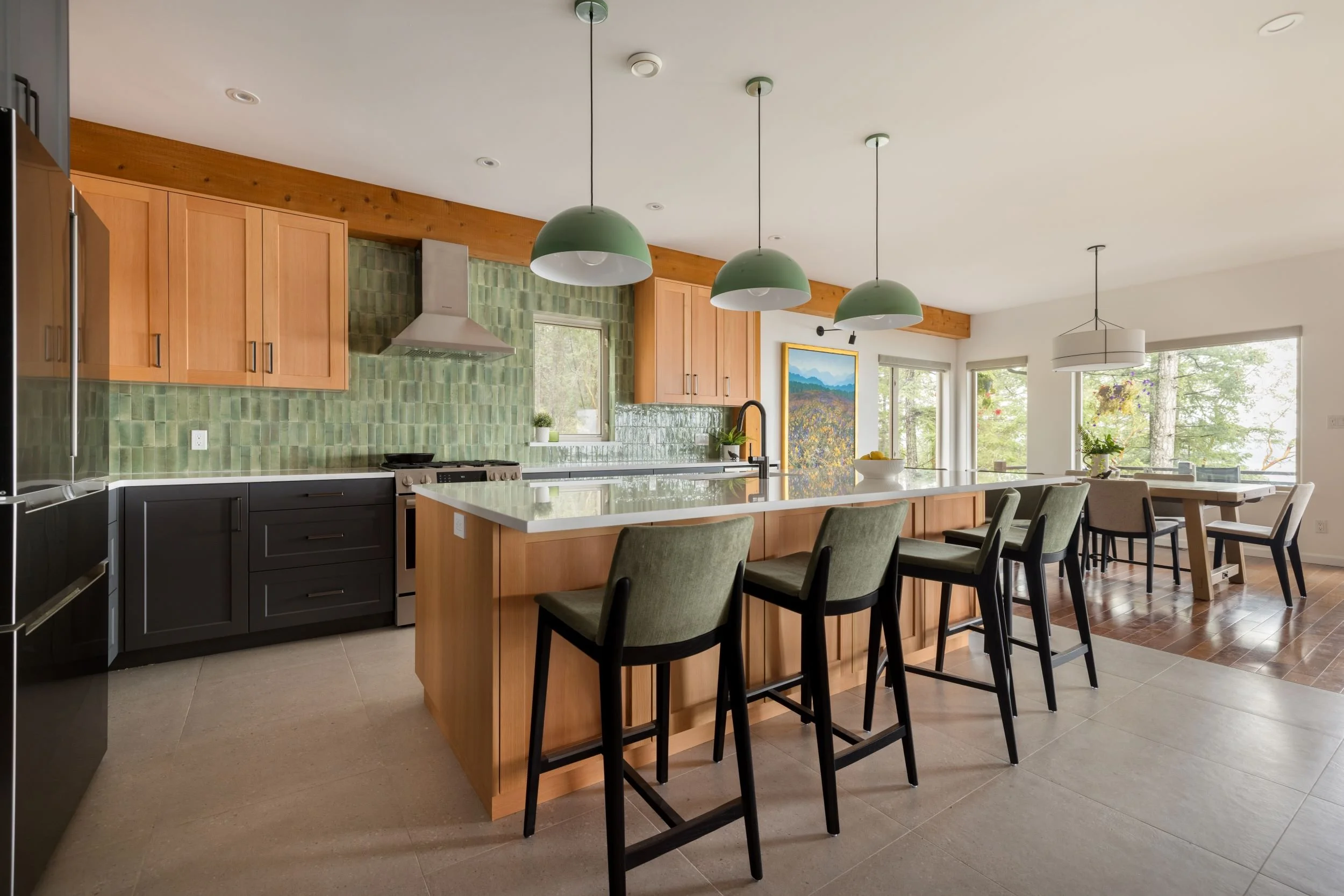 Modern kitchen with green and black cabinets, wooden accents, and a large island with bar stools, overlooking a dining area with large windows and outdoor view.