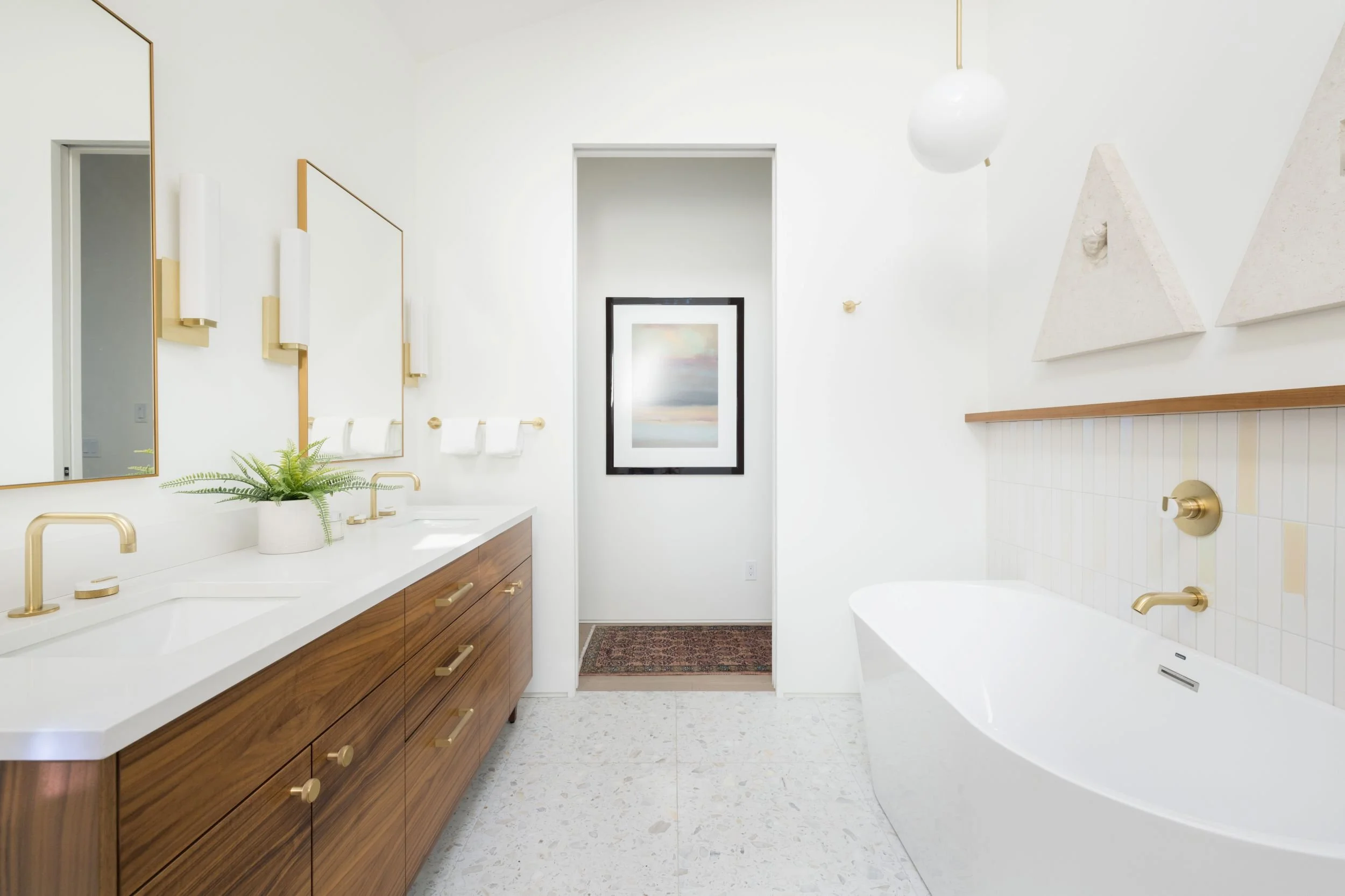 Modern bathroom with white walls, a large wooden vanity with gold fixtures, a white bathtub, and minimalistic decor including wall art and indoor plants.