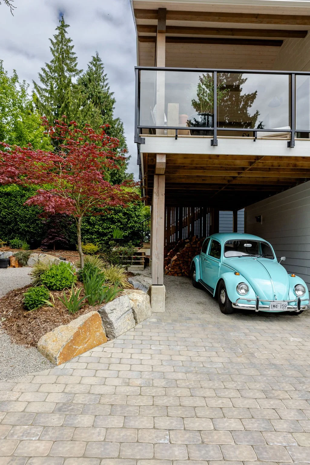 A turquoise vintage Volkswagen Beetle parked in a driveway beneath a two-story house with a balcony, woodpile, and lush greenery including a red-leafed tree.