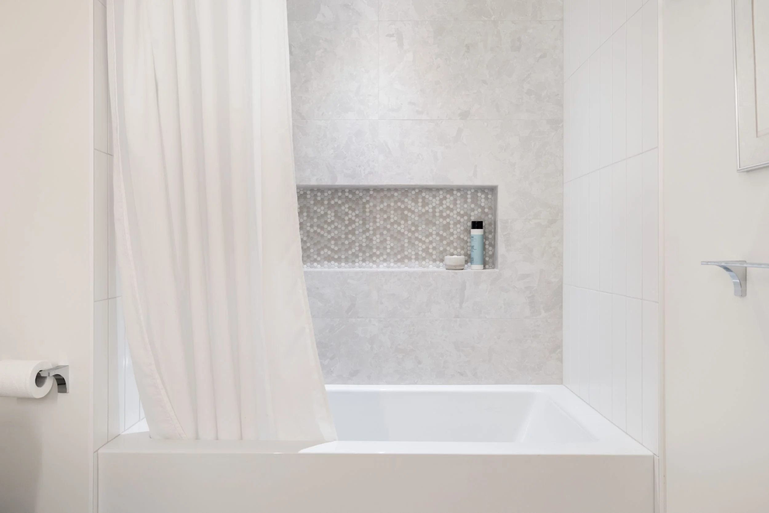 A modern bathroom with a white bathtub, white shower curtain, and light gray tiled walls featuring a built-in niche with toiletries.