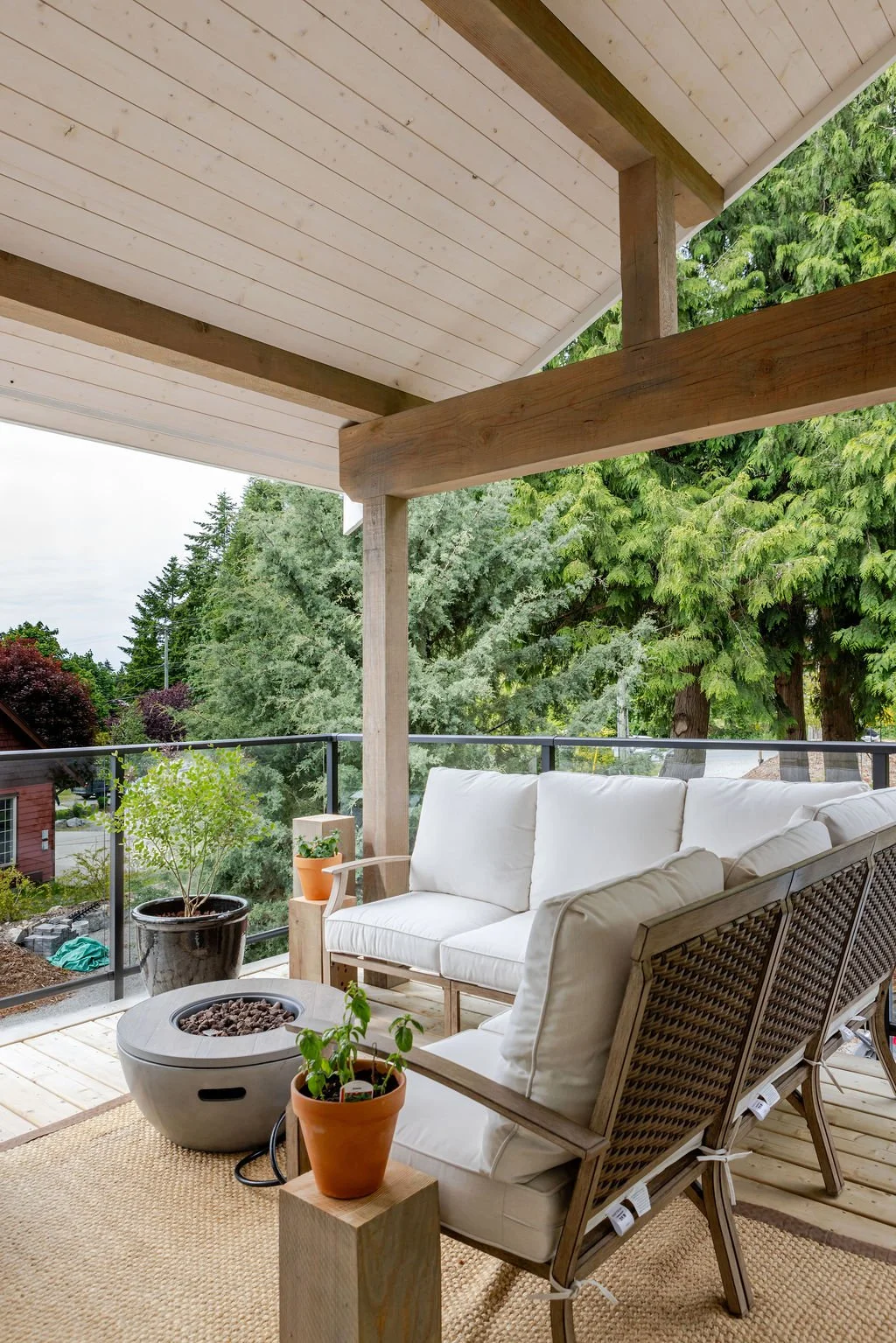 Outdoor balcony with white cushioned furniture, potted plants, and a fire pit, overlooking trees and neighboring houses.