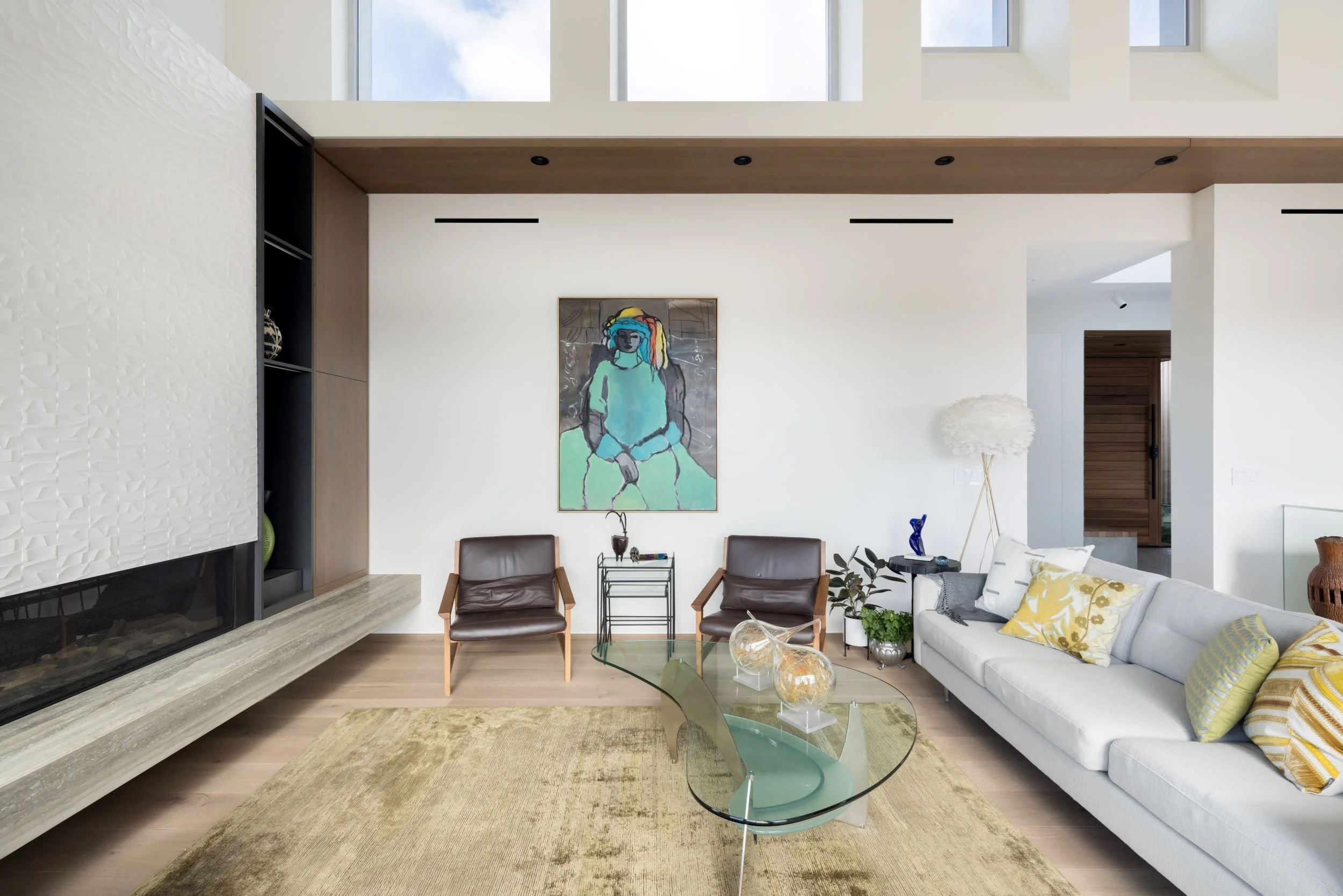 Modern living room with white walls, large windows, a white sofa with yellow and white pillows, a glass coffee table, two brown chairs, a colorful abstract portrait, and decorative items including a white feather lamp, plants, and vases.