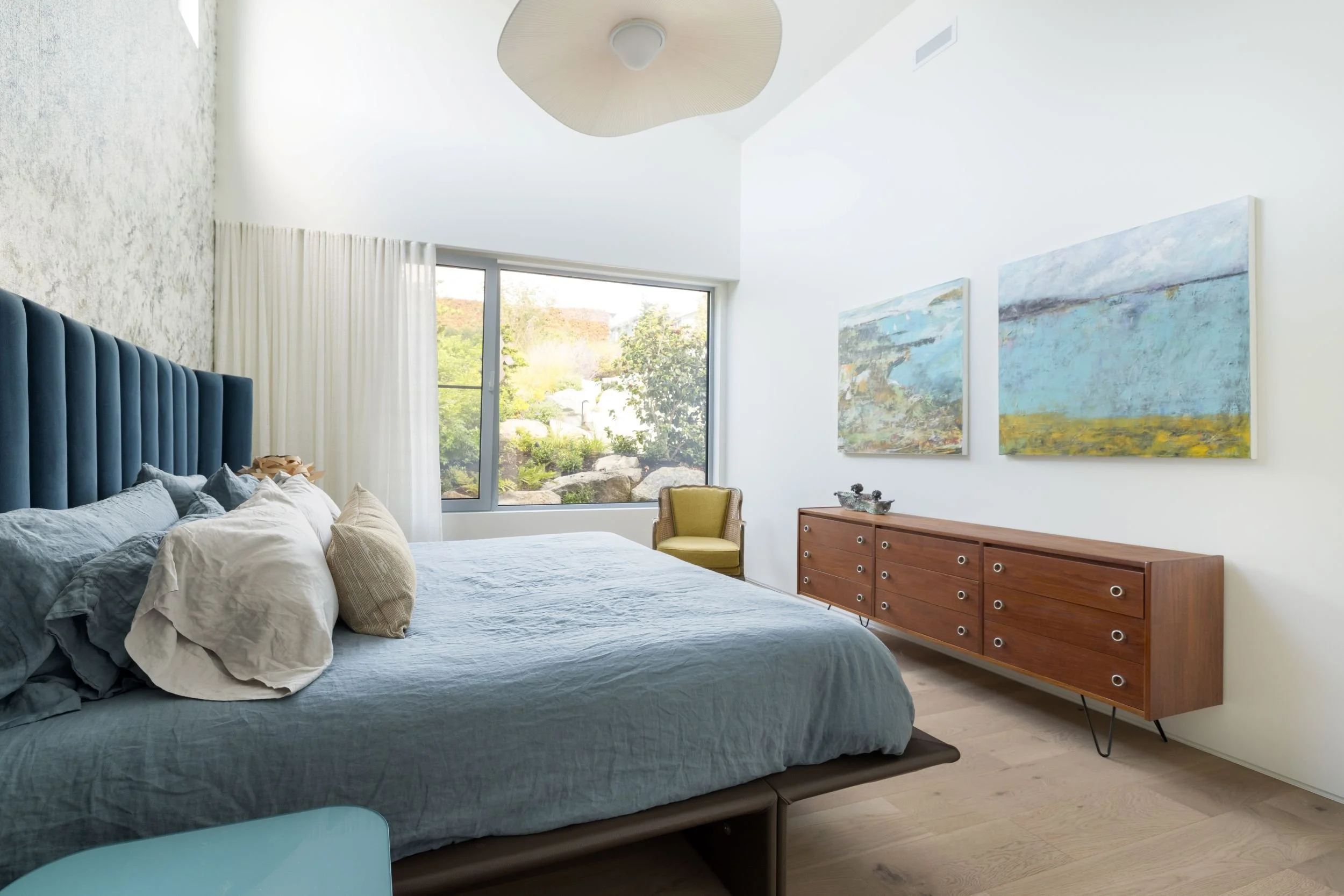 A bedroom with a large bed with blue and gray bedding, a wooden sideboard, artwork on the wall, a window showing a garden, and a yellow armchair.