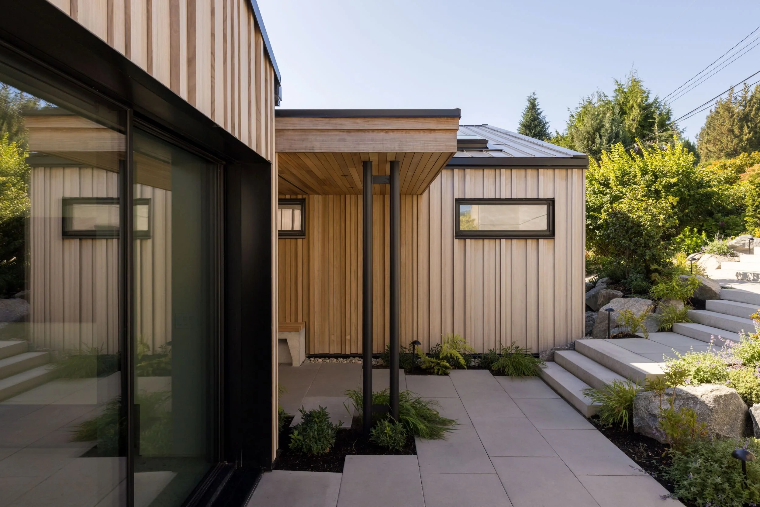 Modern house exterior with wood siding, black-framed windows, concrete steps, and landscaped garden with plants and rocks.