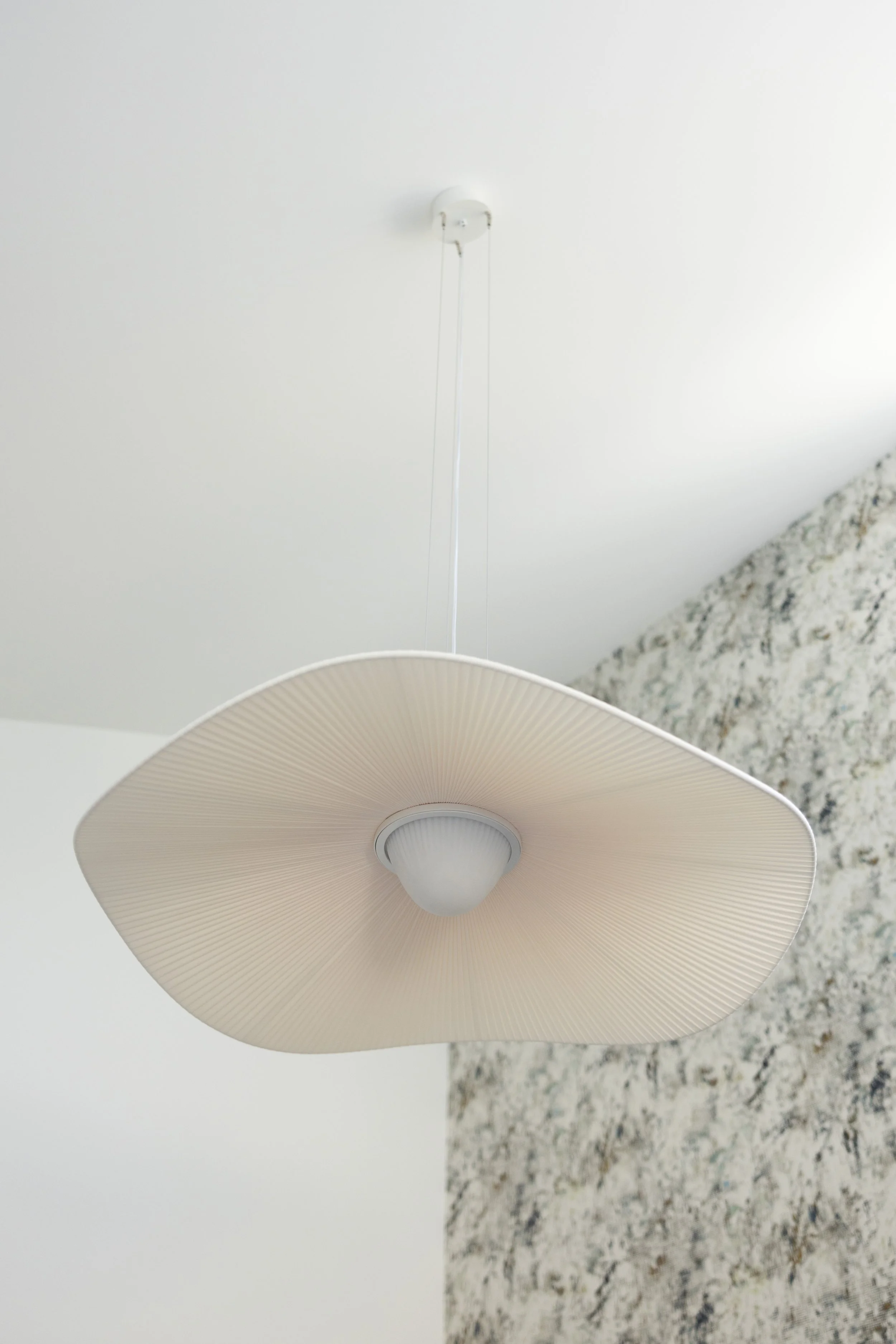 A modern ceiling light fixture with a large, beige, pleated lampshade hanging from a white ceiling, positioned above a textured wall.