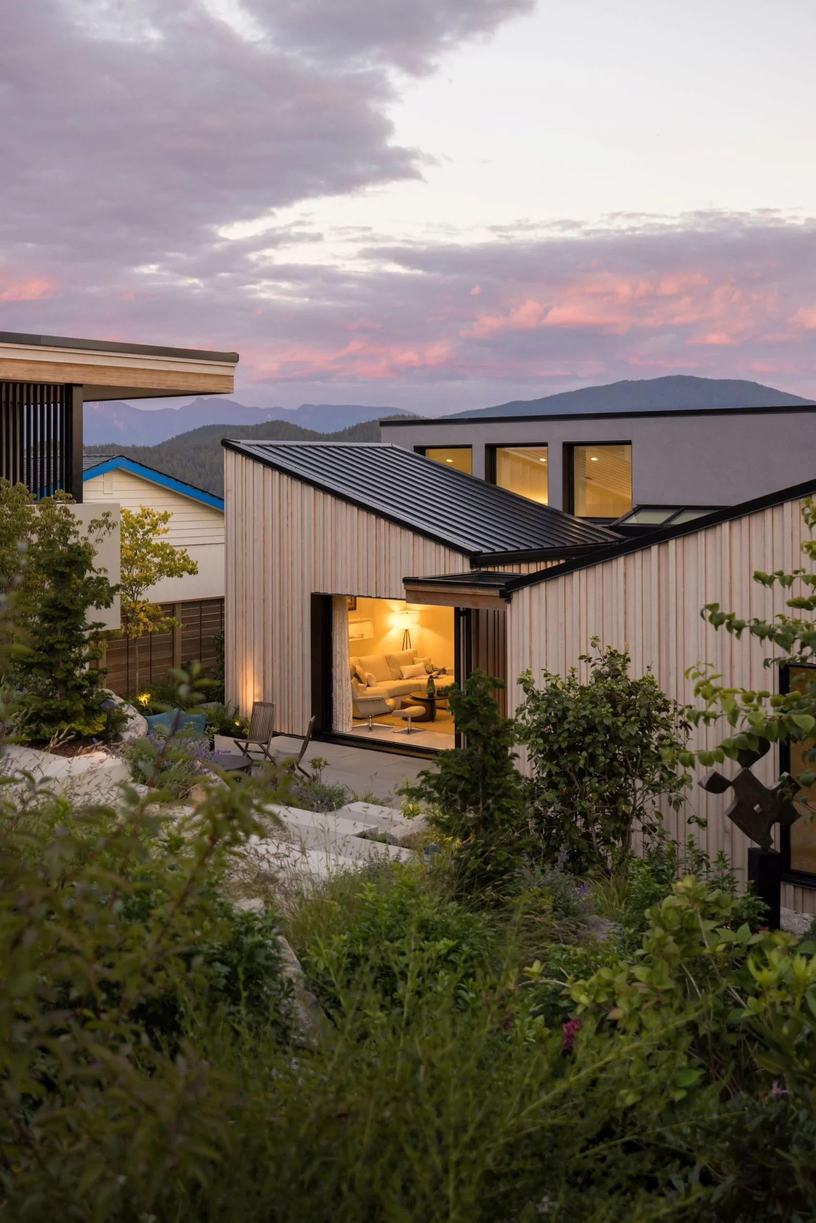 A modern residential house with a lit interior, wooden exterior, set against a mountainous backdrop during dusk, with a garden in the foreground.