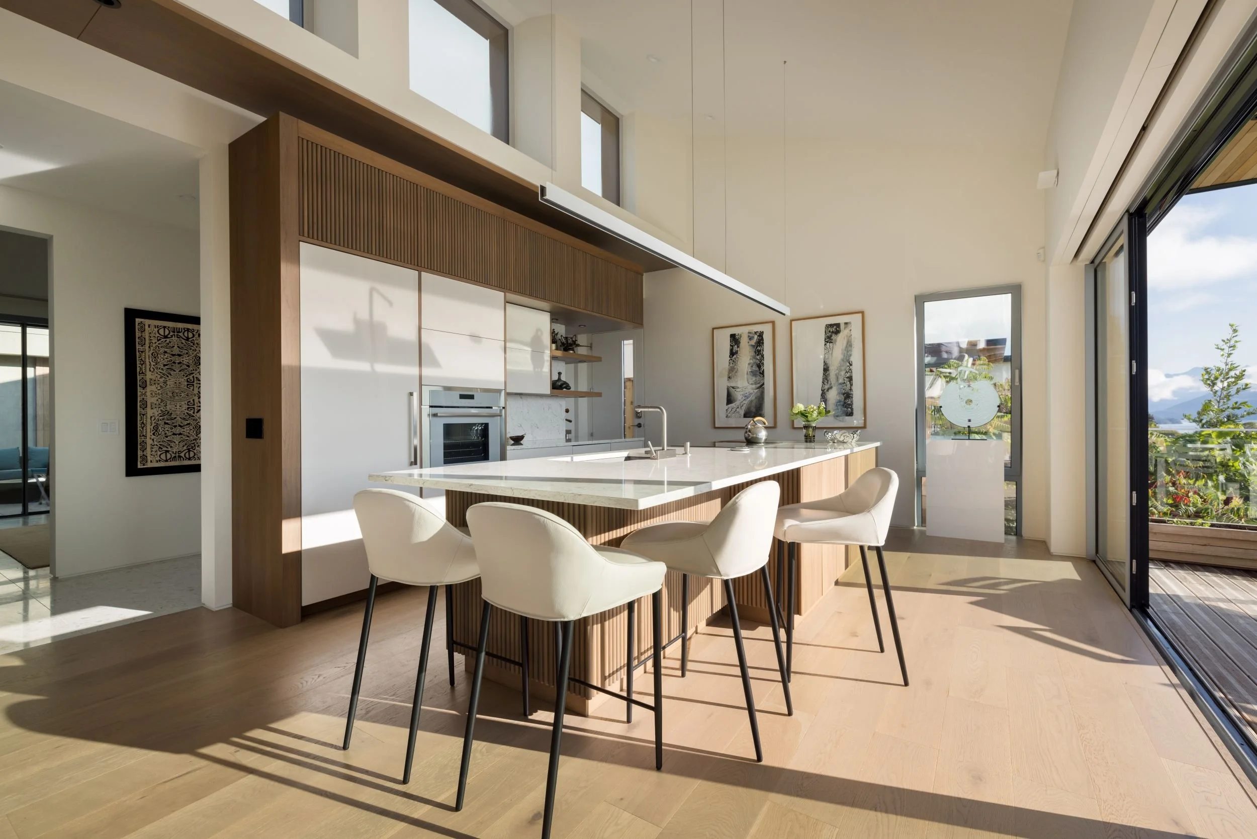 Modern kitchen with white island and four cream-colored chairs, wooden accents, large windows and glass door leading to an outdoor patio with a landscape view.
