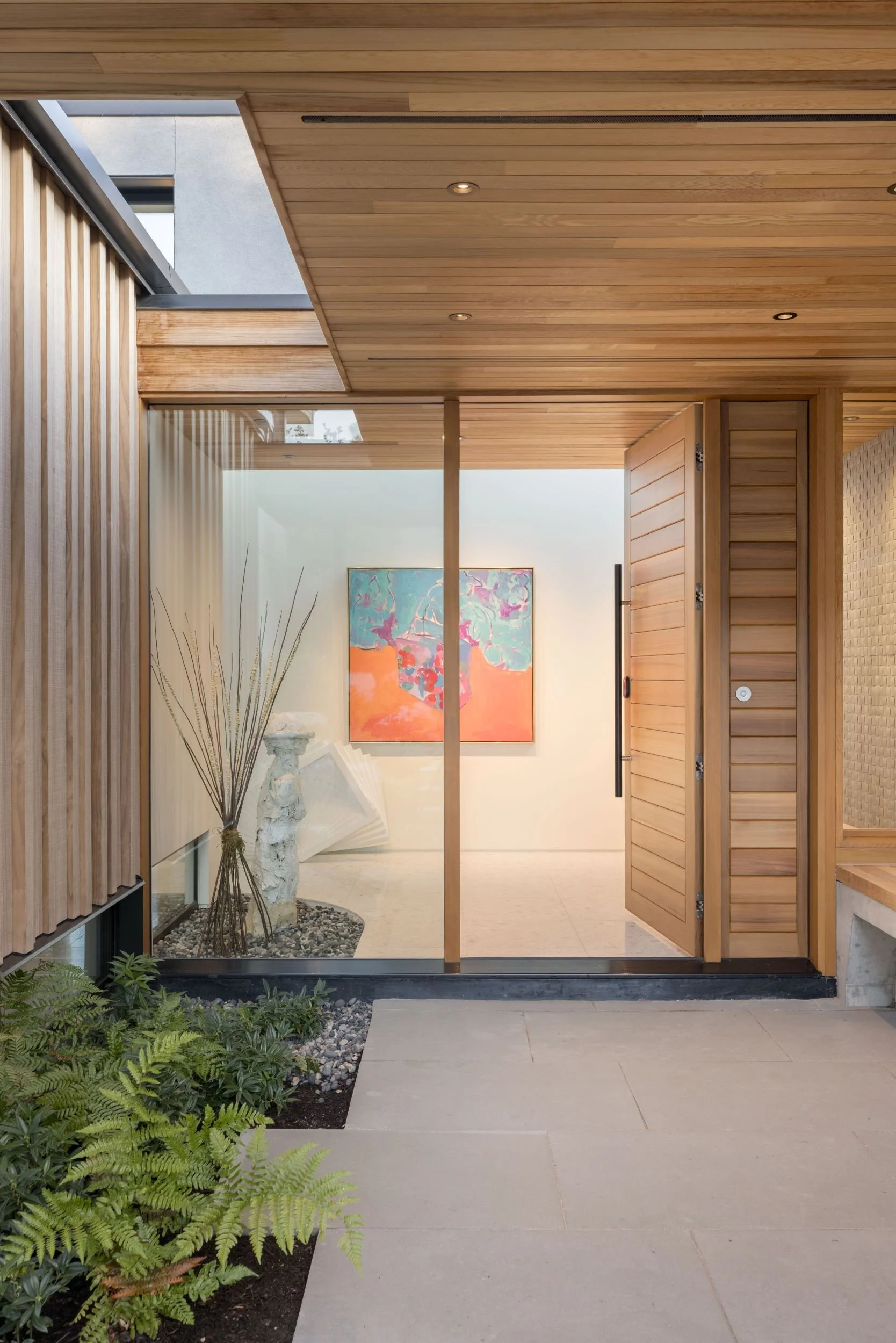 Modern house entrance with wooden sliding doors, a small outdoor garden with ferns, and an interior hallway featuring abstract artwork and decorative sculptures.