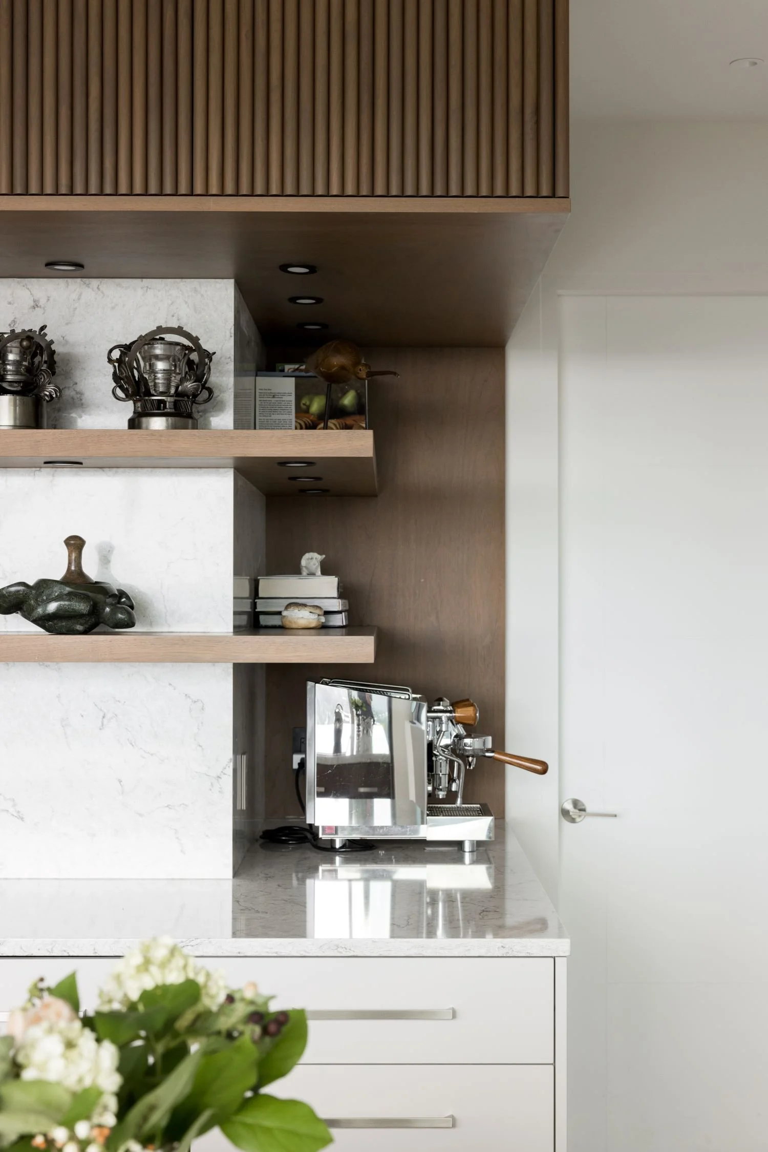 Modern kitchen with white marble countertops, wooden shelves, and a silver espresso machine on the counter.