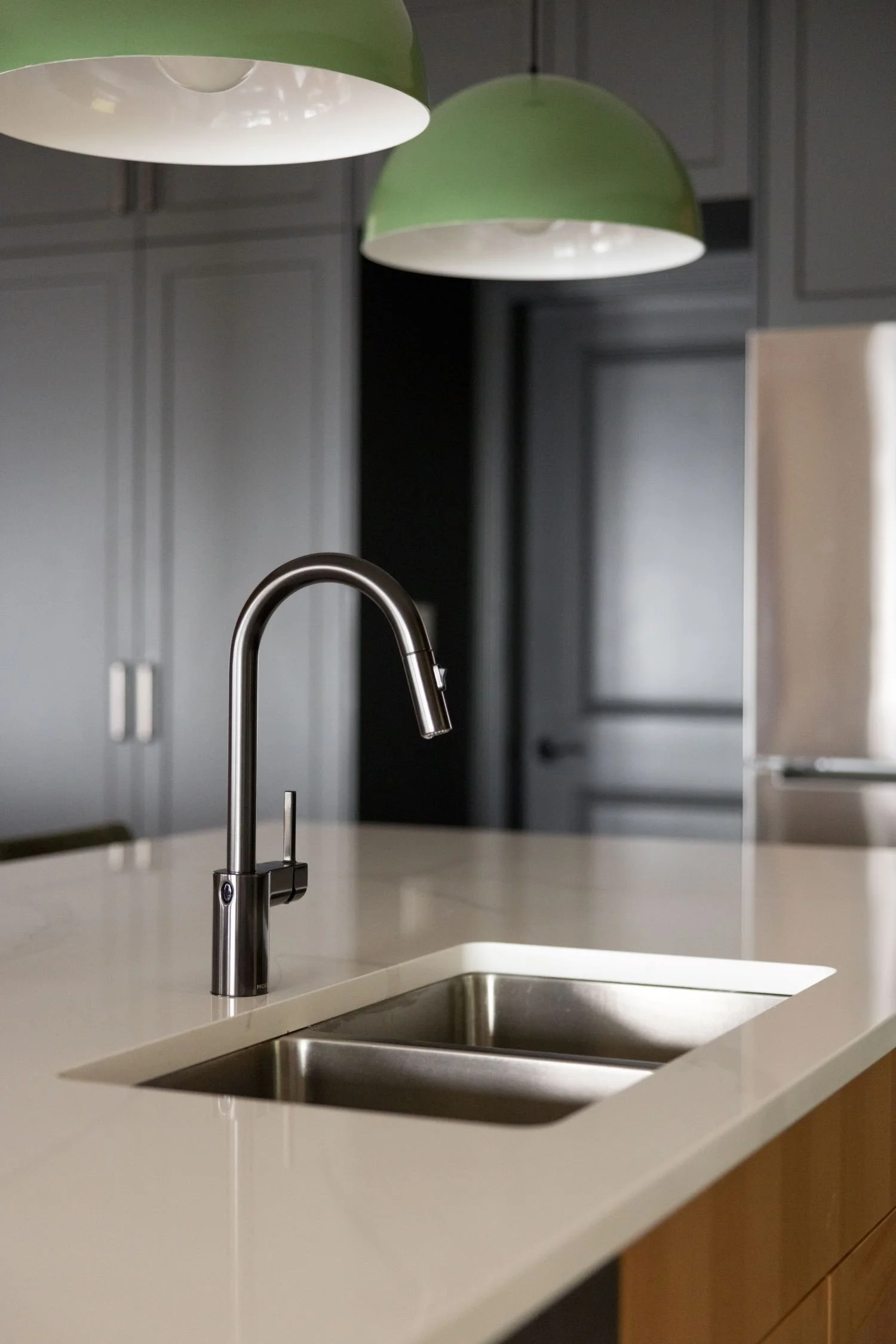 Close-up of a modern kitchen sink with a curved stainless steel faucet on a white countertop, with dark gray cabinetry and two green pendant lights hanging above.