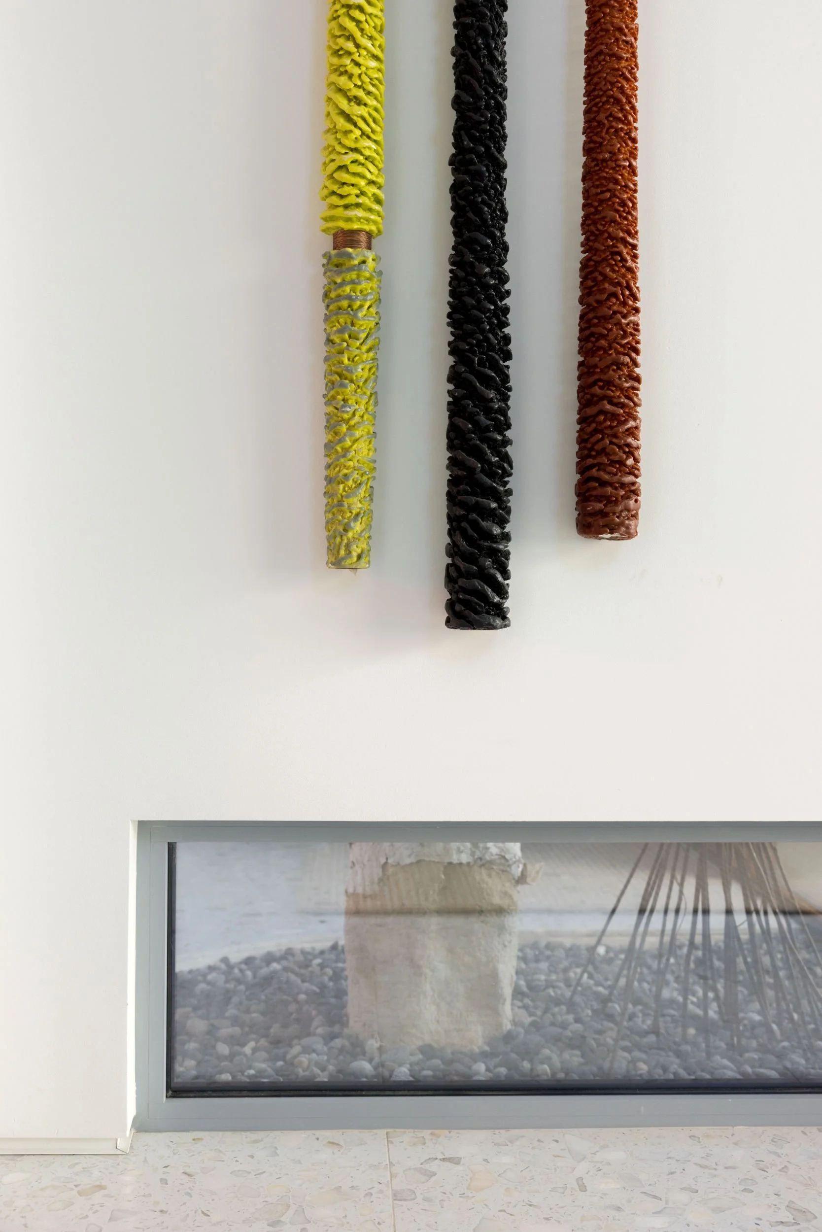 Three textured vertical art sculptures in yellow, black, and red on a white wall above a low window with a view of a stone feature and decorative item outside.