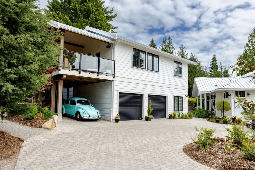 Modern white house with black trim, large windows, and a balcony with glass railing. An open garage with a vintage blue car parked inside. Paved driveway with potted plants and surrounding greenery. Clear sky with scattered clouds.