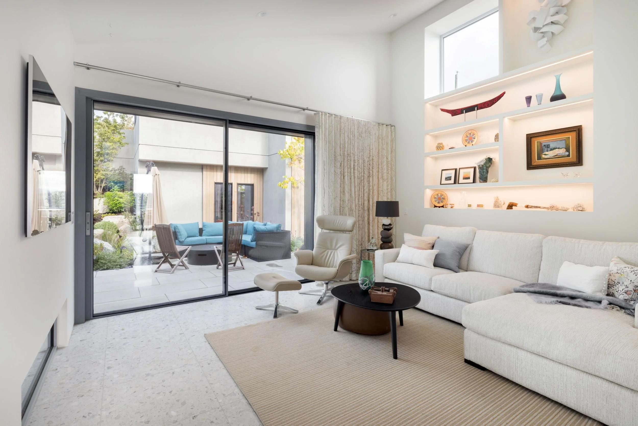 Living room with white sectional sofa, armchair, black coffee table, built-in wall shelves with decor, sliding glass door to outdoor patio with seating area.