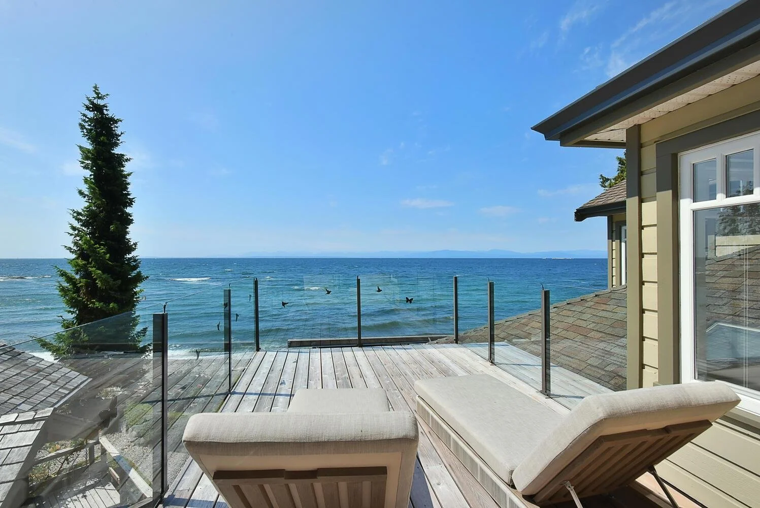 A wooden deck with two lounge chairs overlooking the ocean with a glass railing, a house to the right, a tall tree to the left, and a clear blue sky in the background.