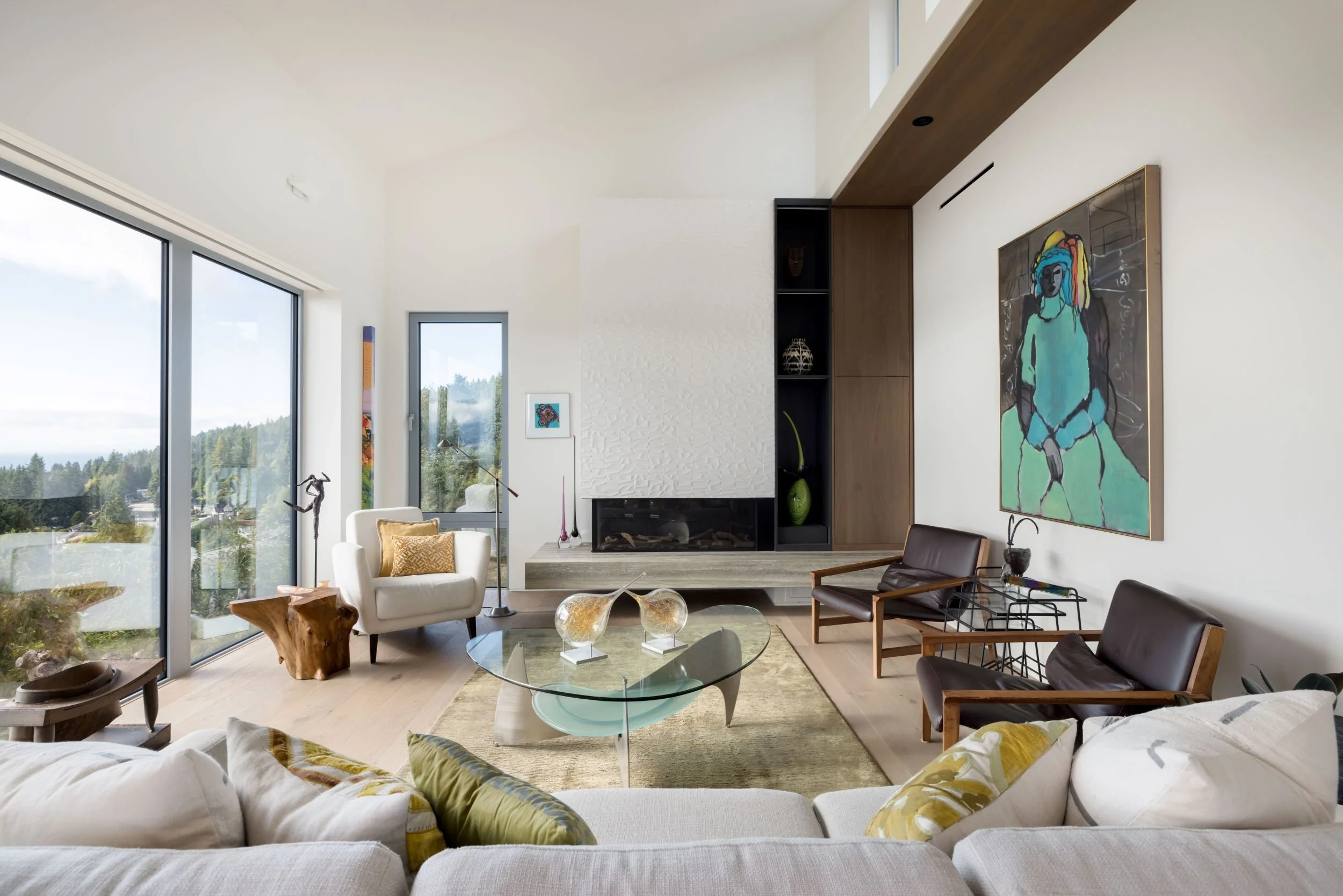 Modern living room with large glass windows, white walls, wooden floors, contemporary furniture, a fireplace, and colorful artwork.