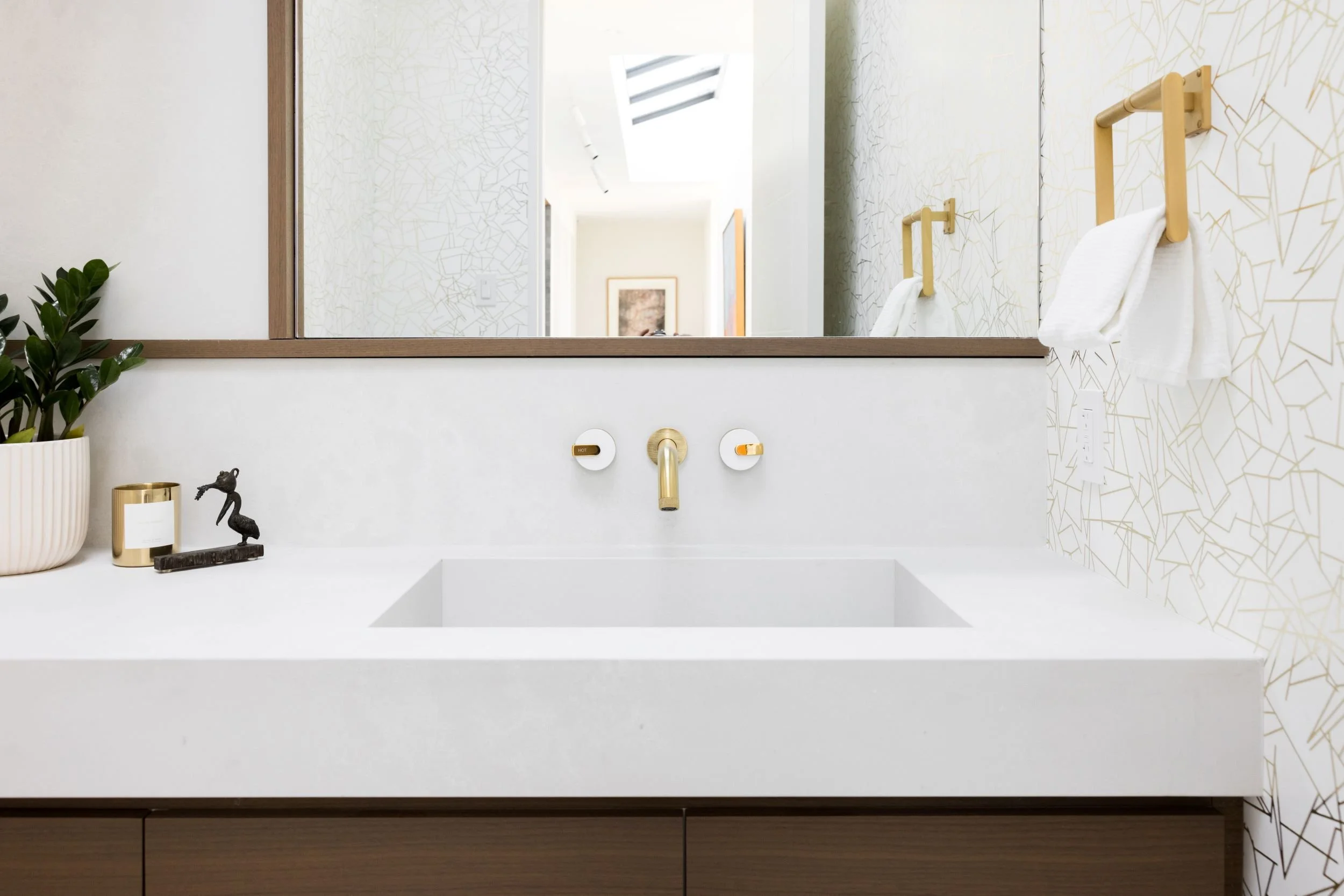Modern bathroom vanity with white countertop, gold fixtures, a large mirror, and decorative items including a potted plant, a candle, and a small sculpture of a horse.