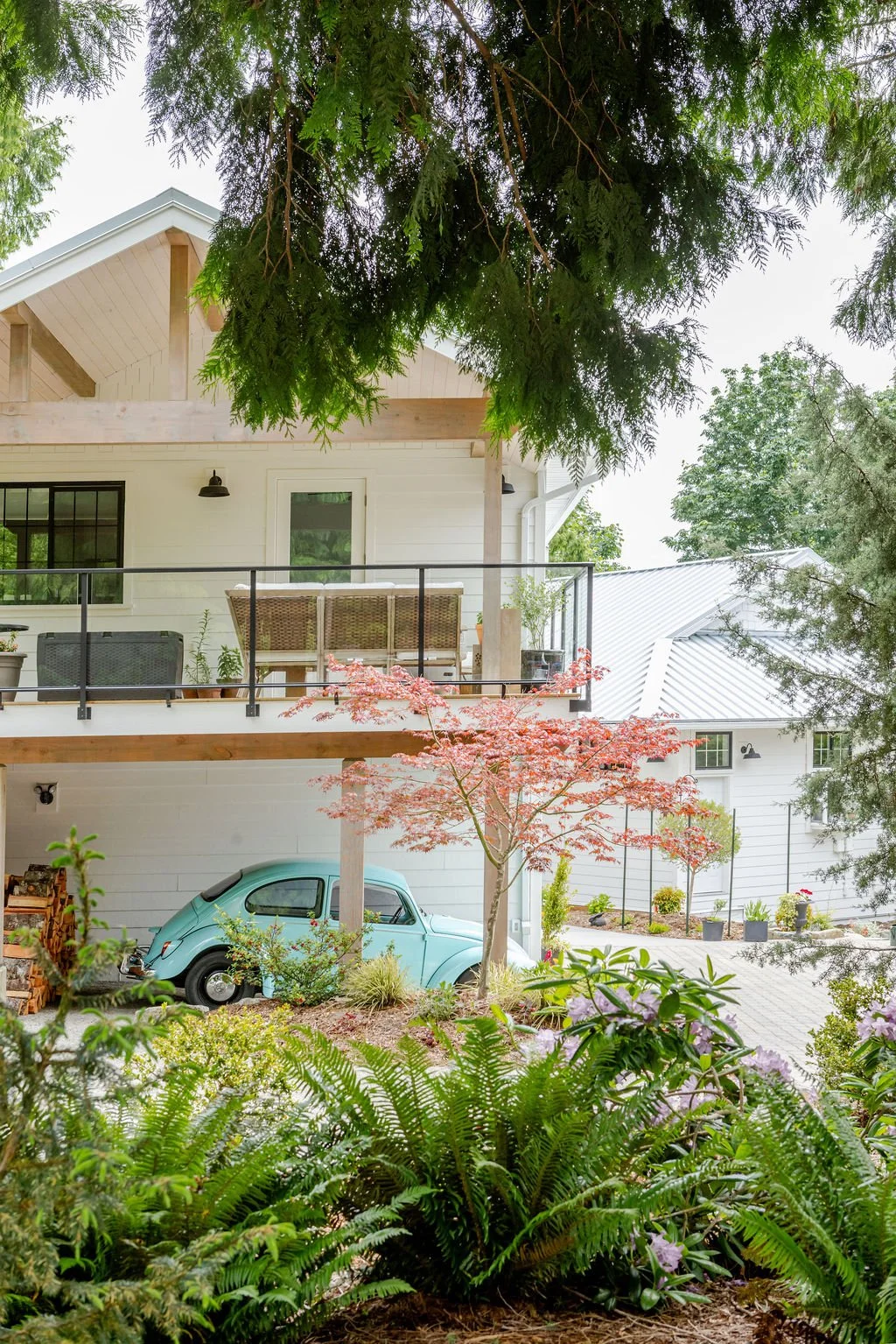 View of a house with a front porch, balcony, and garden with green trees, plants, and a vintage blue car parked underneath the balcony.