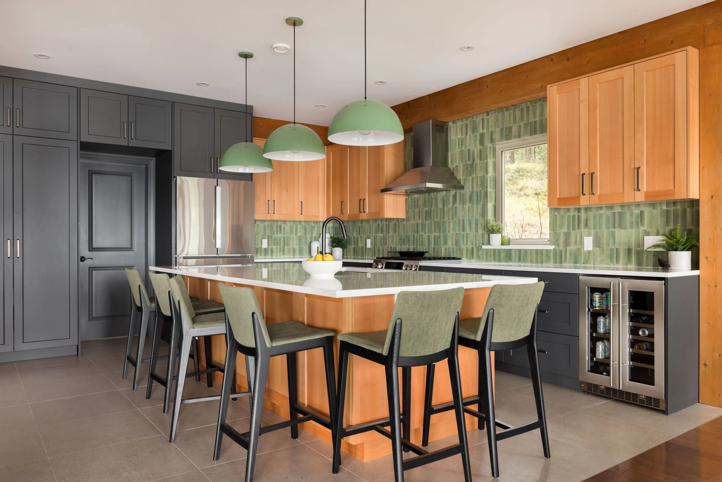 Modern kitchen with gray cabinets, wooden accents, green backsplash tiles, and an with green chairs. Pendant lights hang above the island, and there is a window with a view of greenery outside.