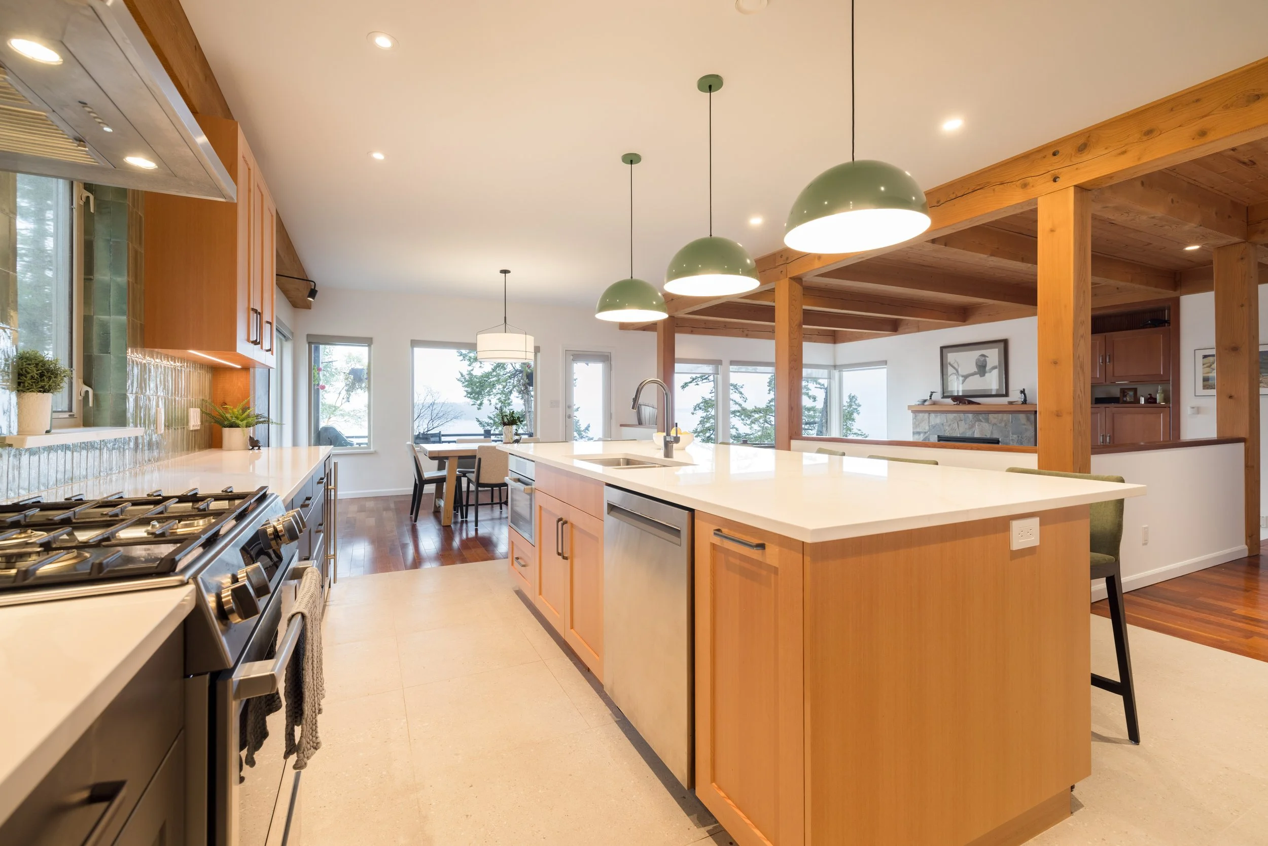 Modern kitchen with wooden cabinetry, a large white island, pendant lights, and dining area with windows overlooking trees.