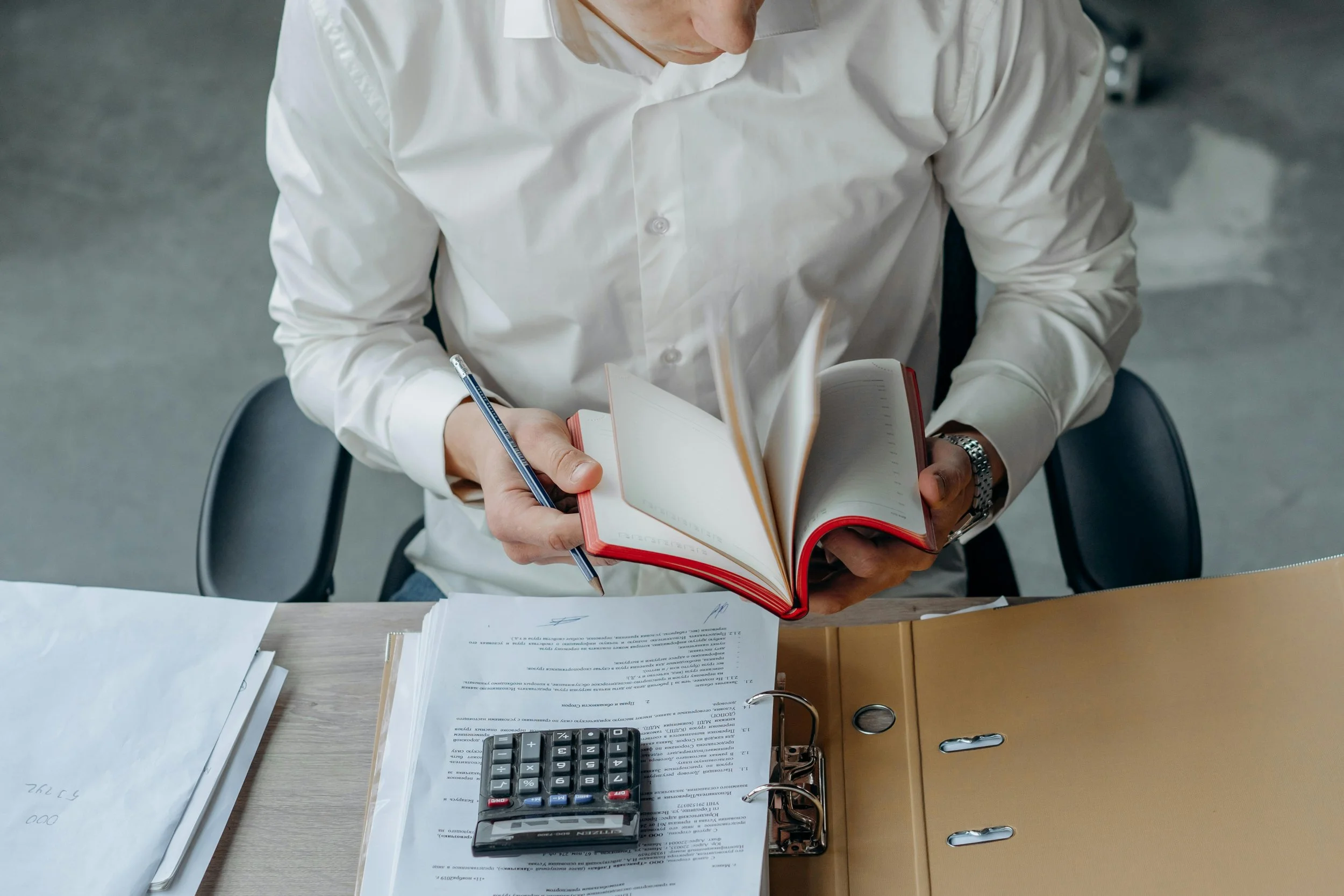A person in a white shirt sitting at a desk, reading a book, with papers, a calculator, and a pen on the desk.