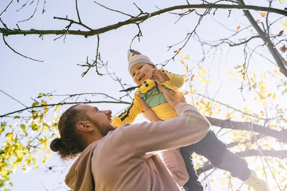 Man lifting a child wearing a winter coat into the air, with a sunny sky and bare tree behind them.