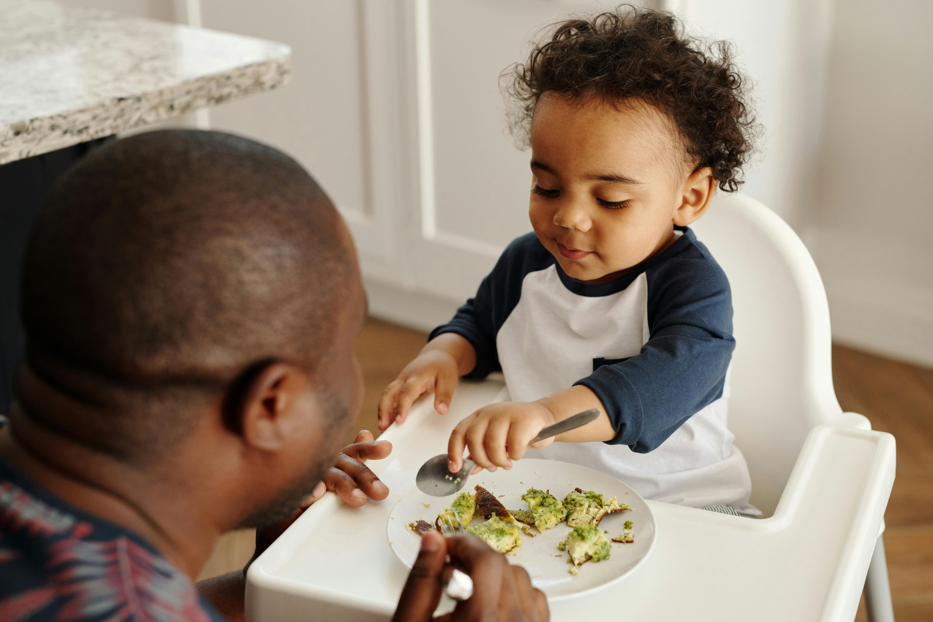 Toddler seated in a high chair eating with a caregiver nearby.