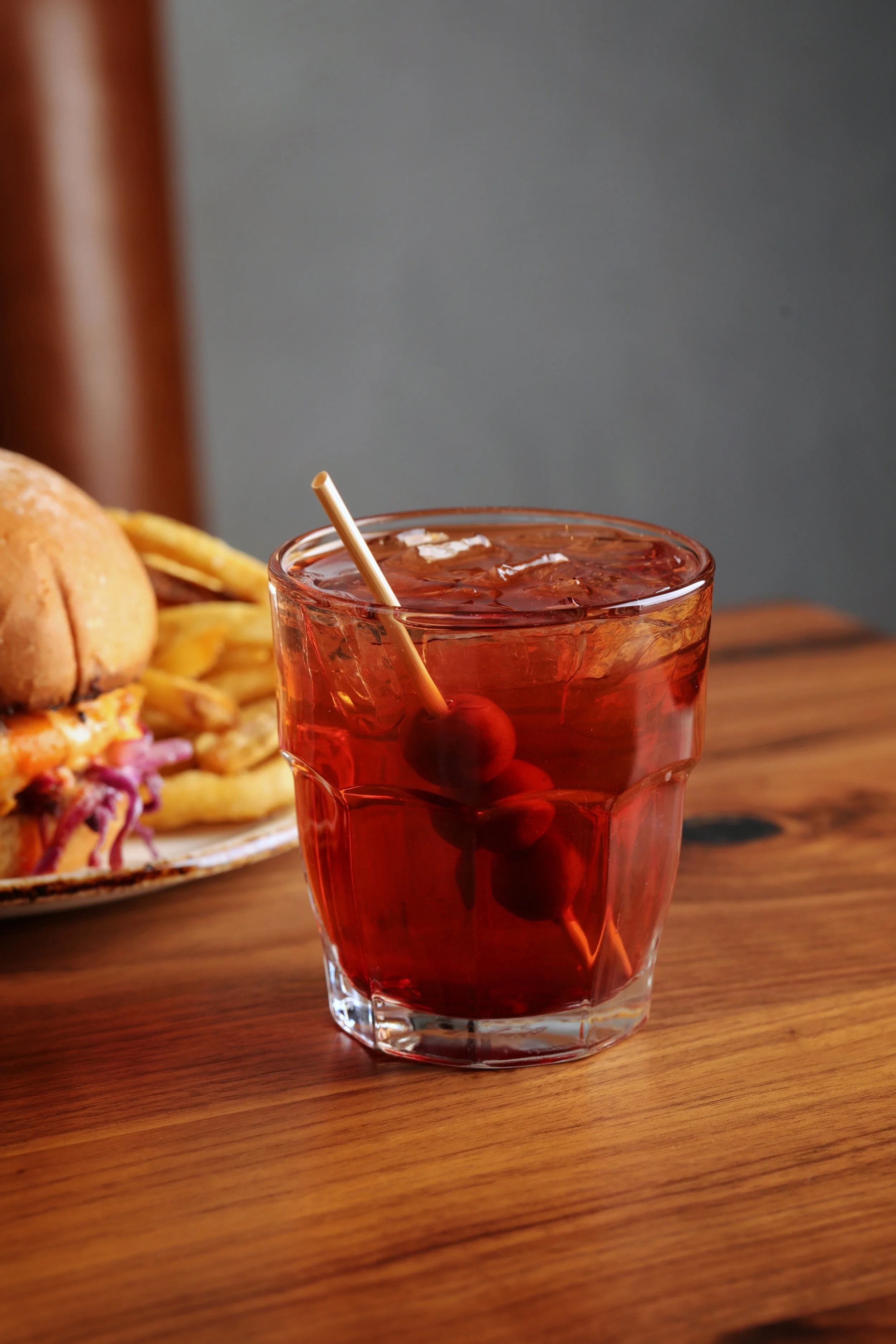 A glass of iced cocktail with skewered olives and a burger with fries in the background on a wooden table.