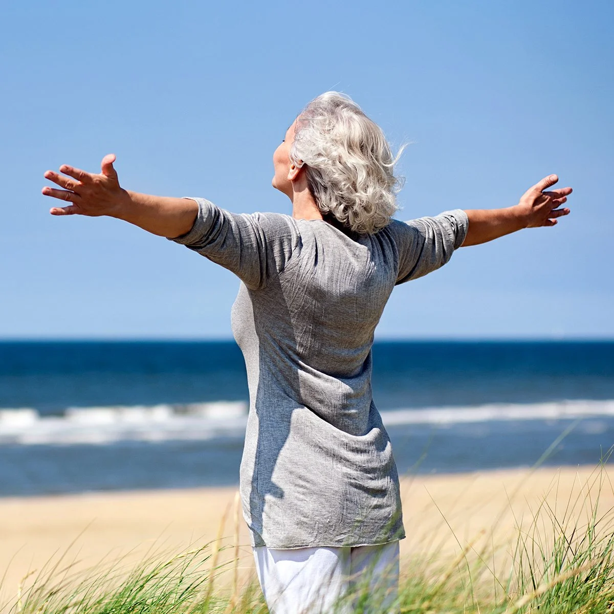 An older woman with gray, curly hair standing on a grassy beach with her arms outstretched, facing the sky, with ocean waves and a clear blue sky in the background.