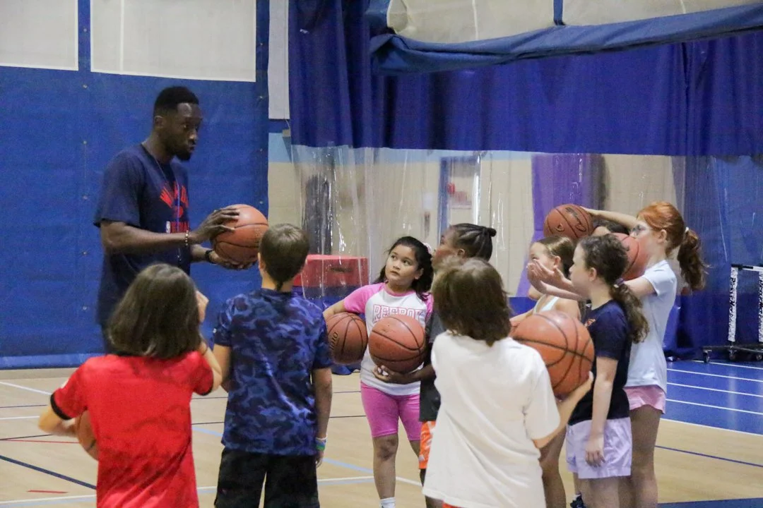 A basketball coach instructs a group of children holding basketballs in a gymnasium.