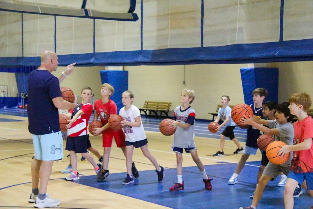 A coach instructs young children during a basketball practice in an indoor gym, with the children holding basketballs.
