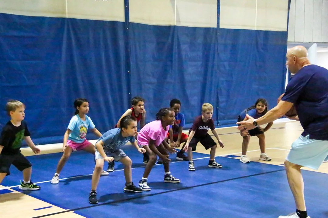 A coach demonstrates a stretching or warm-up exercise to a group of children in a gymnasium.