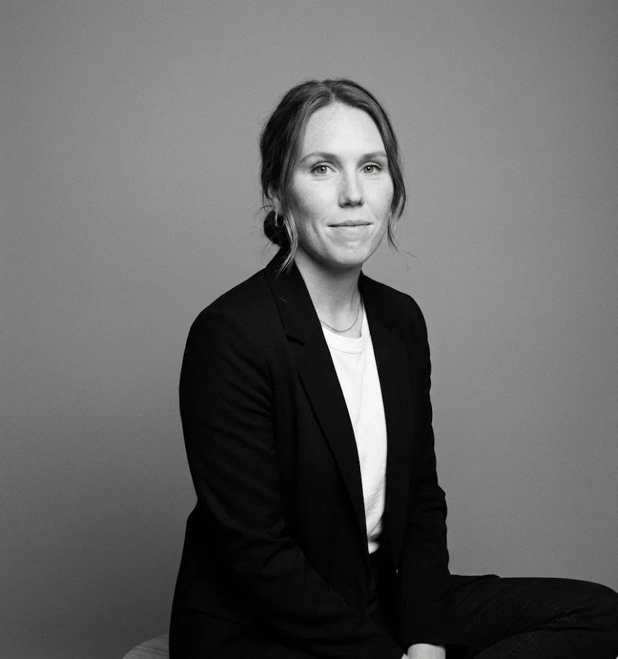 Black and white portrait of a woman sitting against a plain background, wearing a dark blazer over a light shirt, with her hair pulled back and a subtle smile.