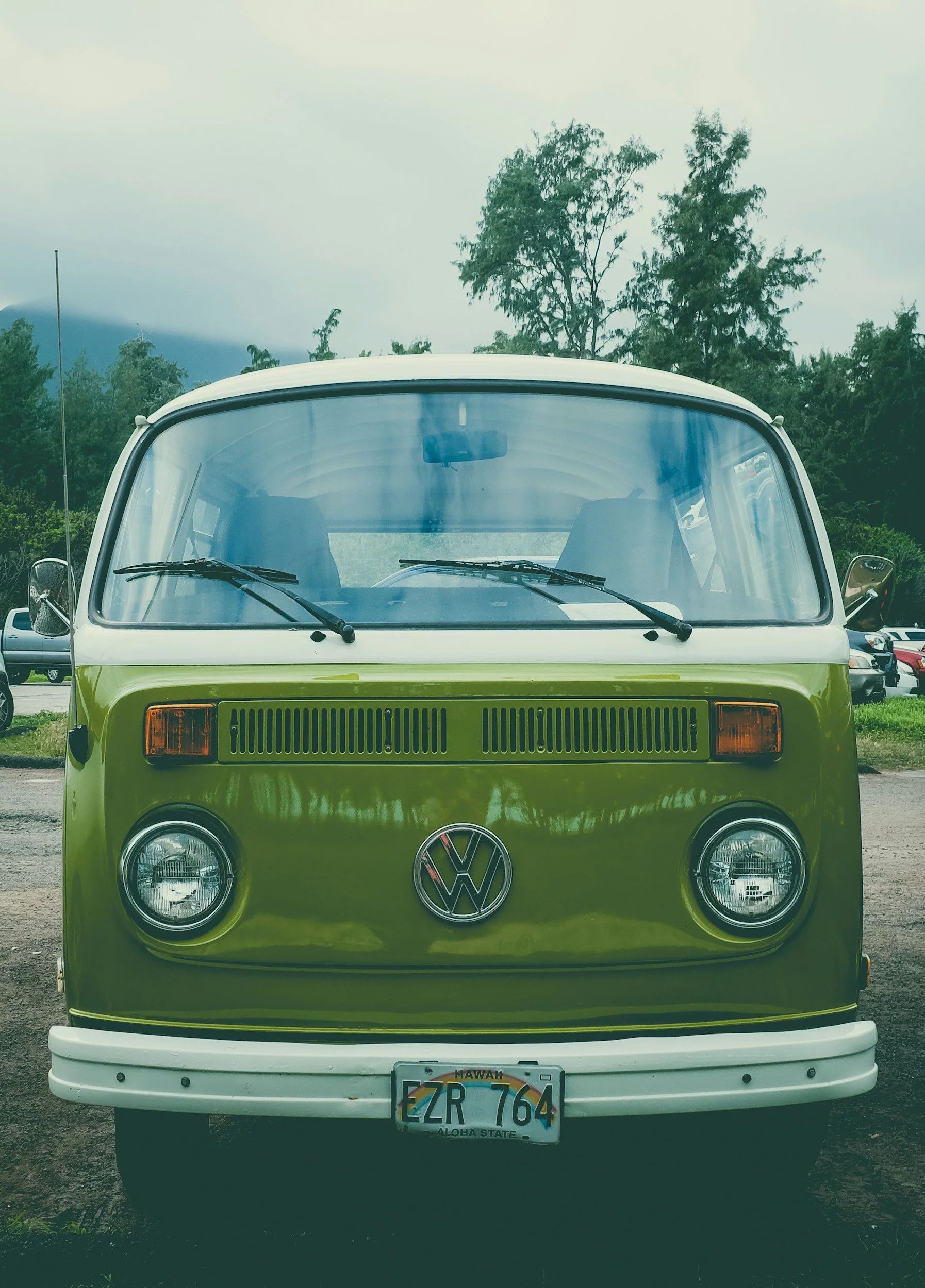 Front view of a vintage lime green Volkswagen camper van with a white roof, parked outdoors on a cloudy day with trees in the background.