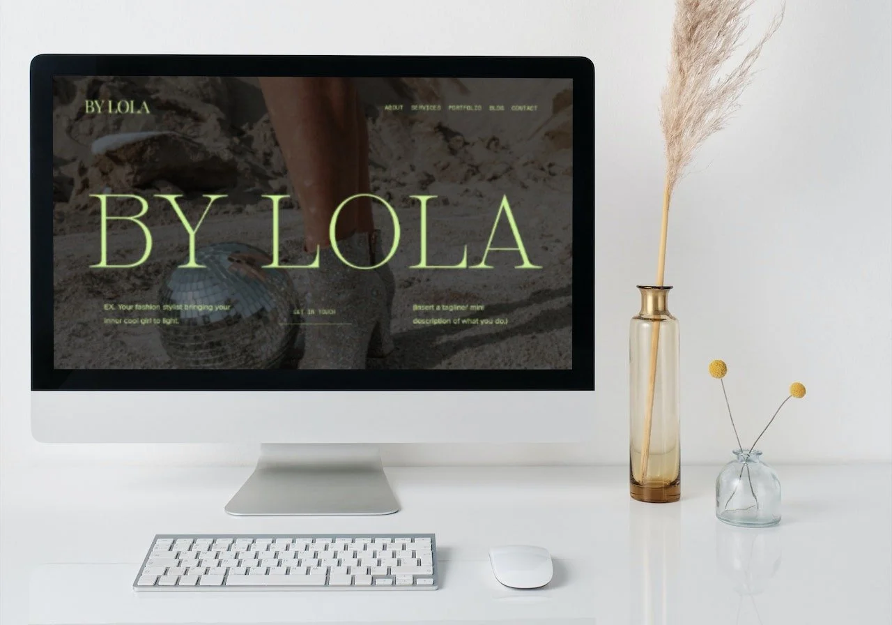 A white desk with an iMac computer displaying a website named BY LOLA, a wireless keyboard, and a mouse. On the right side, there are two glass vases with dried pampas grass and small yellow flowers.