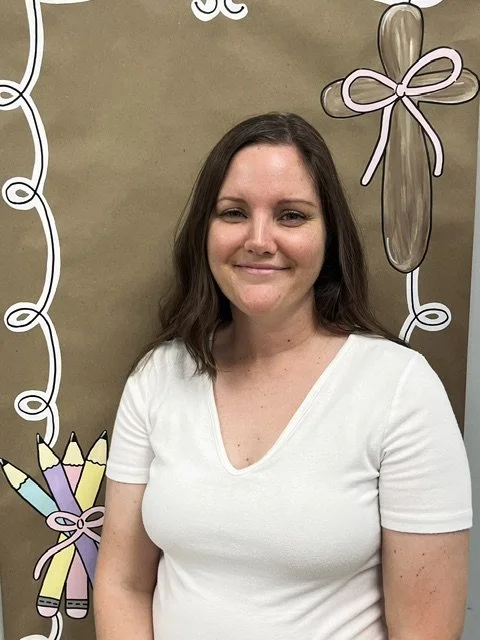 A woman standing outdoors next to a tree with green leaves and a brick wall in the background. She is smiling, wearing glasses, a blue top, a beige cardigan, and a gold necklace.